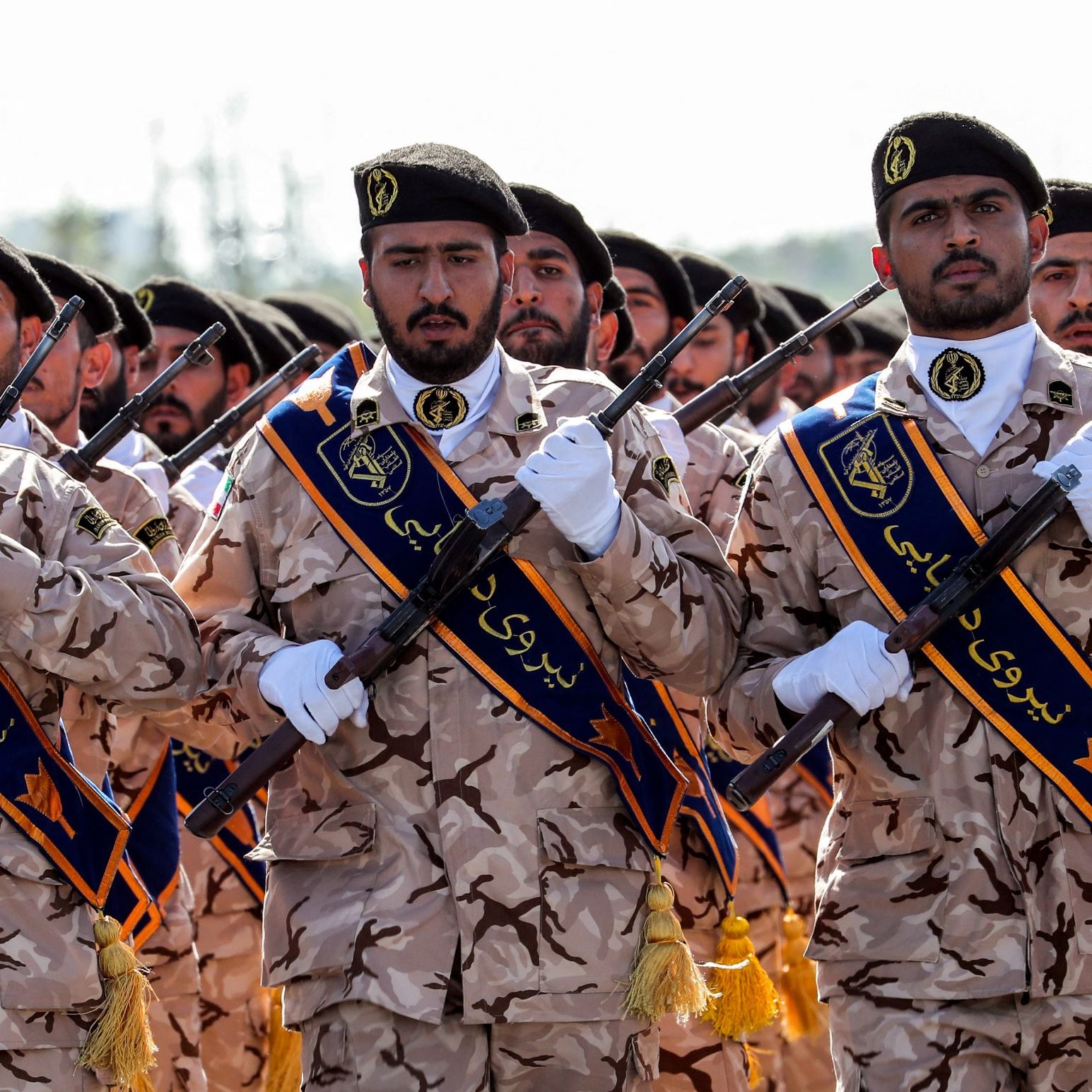 La imagen muestra a un grupo de soldados en un desfile militar. Están vestidos con uniformes de camuflaje y llevan distintivos en sus chaquetas. Los soldados sostienen rifles en posición vertical y parecen estar marchando con solemnidad. El ambiente es solemne y refleja un evento militar con un enfoque en la disciplina y la unidad.