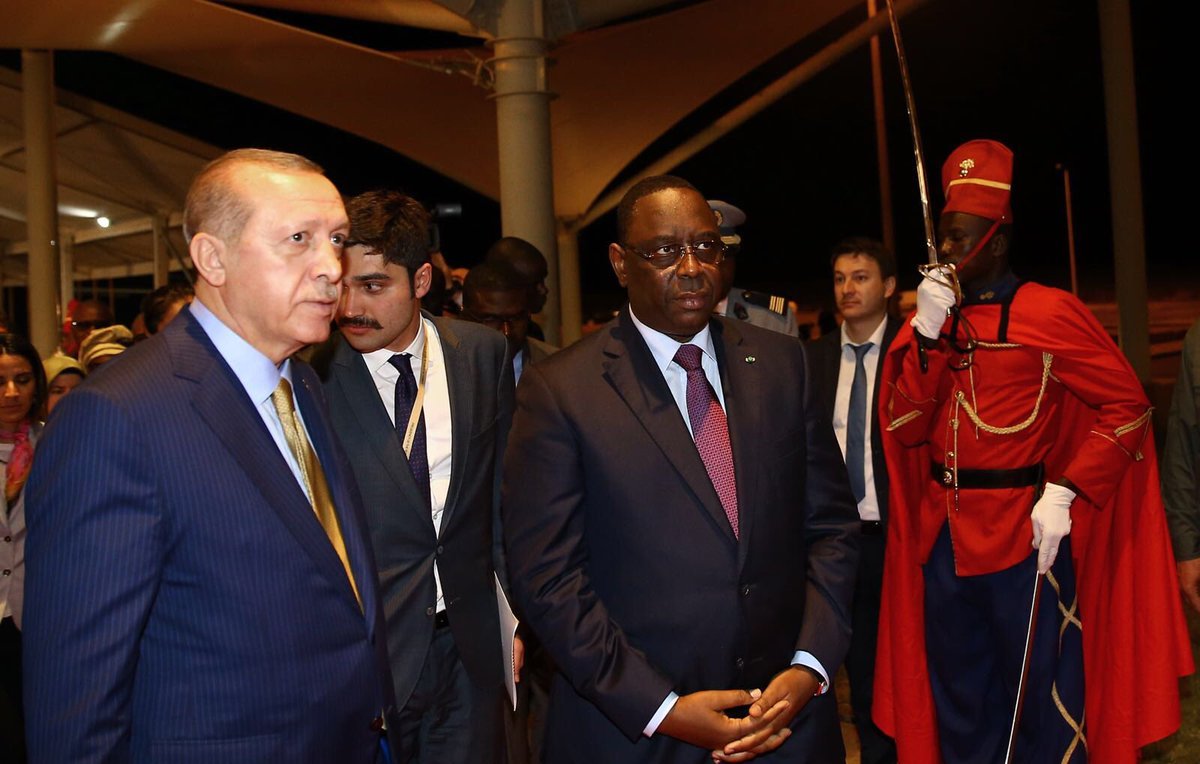 The image shows two prominent figures walking together in a formal setting. One is wearing a dark suit with a light blue striped tie, and the other is dressed in a dark suit with a maroon tie. They appear to be engaged in conversation or deep in thought. In the background, there is a ceremonial guard dressed in a bright red uniform, standing at attention, adding a sense of formality to the scene. The environment looks like an official venue, possibly an airport or a conference center, illuminated by soft lighting.