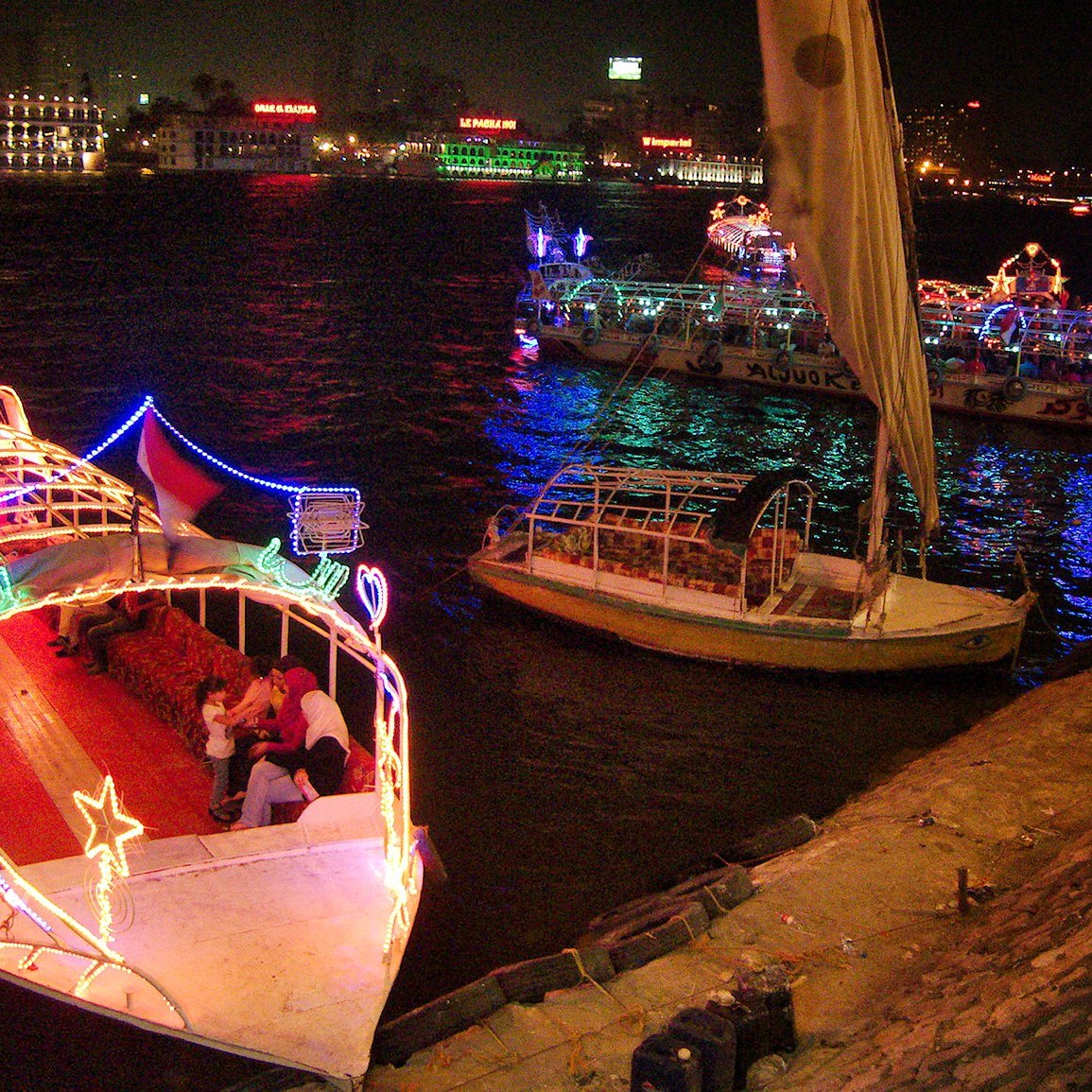 تظهر الصورة مشهدًا ليليًا لنهر، حيث تضيء القوارب بأضواء ملونة وزاهية. يمكن رؤية قوارب أخرى في الخلفية، وبعض الناس يجلسون على ضفاف النهر. تتميز الأجواء بألوان مشرقة تعكس أجواء من الاحتفال والمرح.