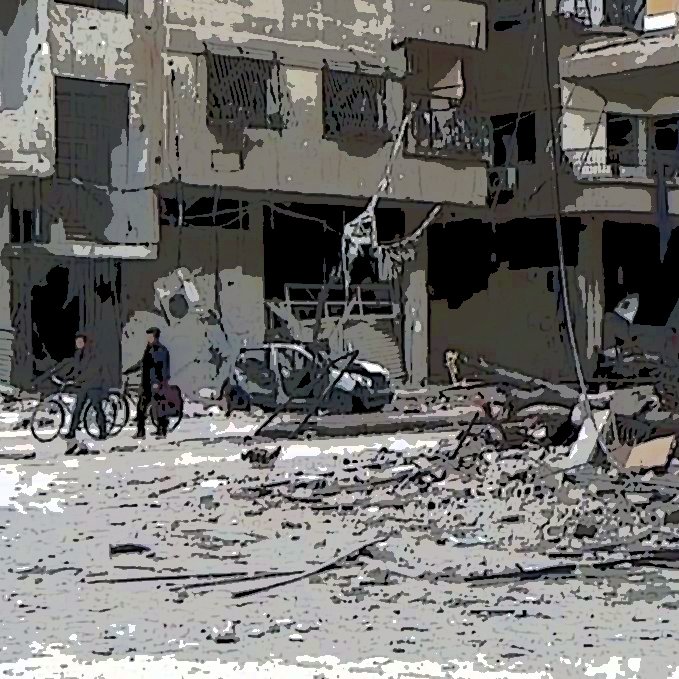 L'image montre une scène de destruction dans une zone urbaine. On y voit des bâtiments endommagés, avec des débris éparpillés sur le sol. Deux personnes marchent au premier plan, tandis que d'autres personnes sont visibles au loin, certaines semblent conduire des vélos. L'ambiance générale est triste et chaotique, illustrant les conséquences dévastatrices d'un conflit.