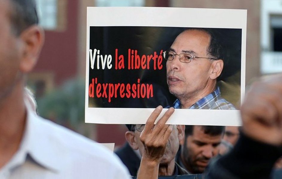 L'image montre une manifestation avec des personnes tenant des pancartes. L'une des pancartes porte le message « Vive la liberté d'expression » en rouge et noir, accompagnée d'une photo d'un homme. L'atmosphère semble engagée et déterminée, reflétant une revendication pour la liberté d'expression. Les manifestants semblent concentrés sur cette cause.