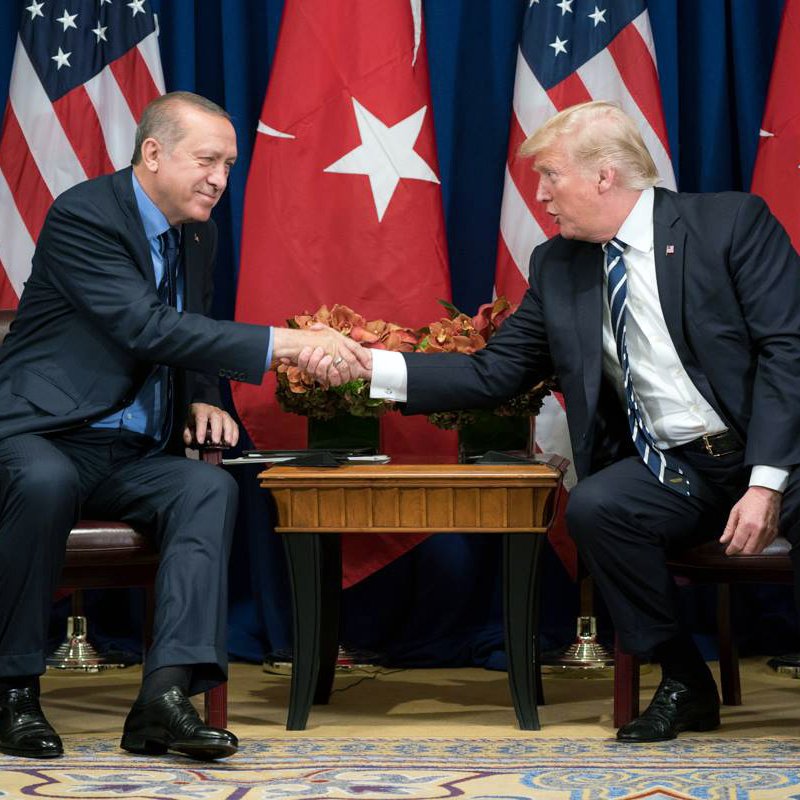 L'image montre deux hommes en costume, engagés dans une poignée de main. Ils sont assis face à face, avec des drapeaux turc et américain en arrière-plan. L'homme à gauche a un visage souriant et une cravate, tandis que l'homme à droite a un regard concentré et une cravate rayée. Un arrangement floral est visible sur la table entre eux. L'ambiance semble être celle d'une rencontre diplomatique.
