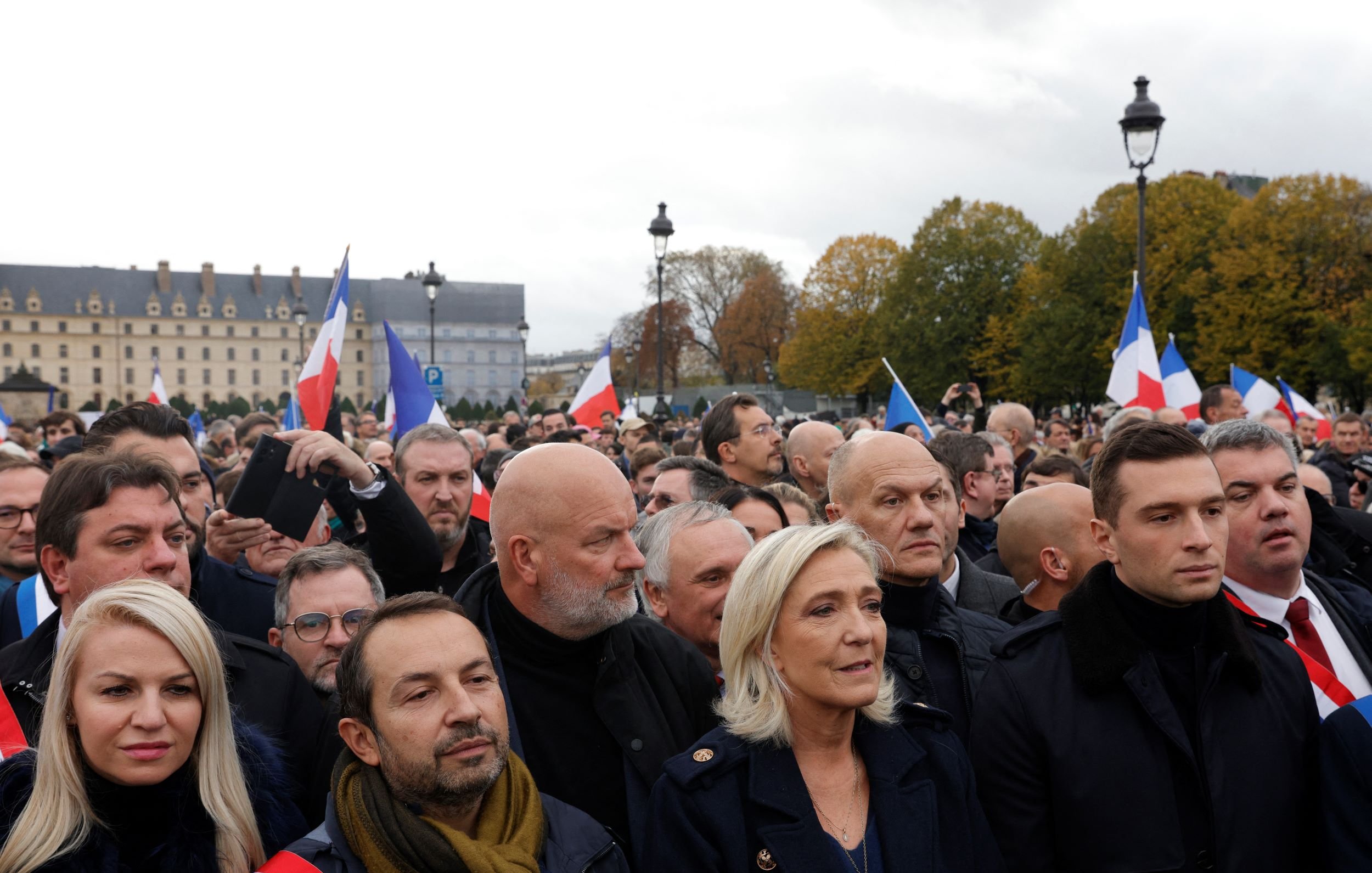 در این تصویر، گروهی از مردم در یک تجمع یا راهپیمایی در فضای باز به نمایش درآمده‌اند. بسیاری از افراد پرچم‌های فرانسه را در دست دارند. در میان جمعیت، افرادی با چهره‌های مختلف دیده می‌شوند که نشان‌دهنده نظرات و احساسات مختلفی هستند. برخی از افراد لباس‌های رسمی و گرم پوشیده‌اند و به نظر می‌رسد موضوع این تجمع سیاسی باشد. آسمان ابری است و فضای عمومی کمی شلوغ و پرتحرک به نظر می‌رسد.