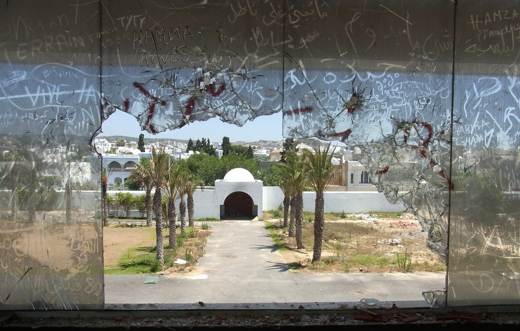 L'image montre une vue à travers un grand trou dans une vitre brisée. On peut voir un paysage urbain avec des palmiers et un édifice en arrière-plan, probablement une entrée ou un portail. Le ciel est dégagé et la scène semble être abandonnée, avec des graffiti visibles sur le verre. L'atmosphère de l'image pourrait évoquer un sentiment de désolation ou d'oubli.