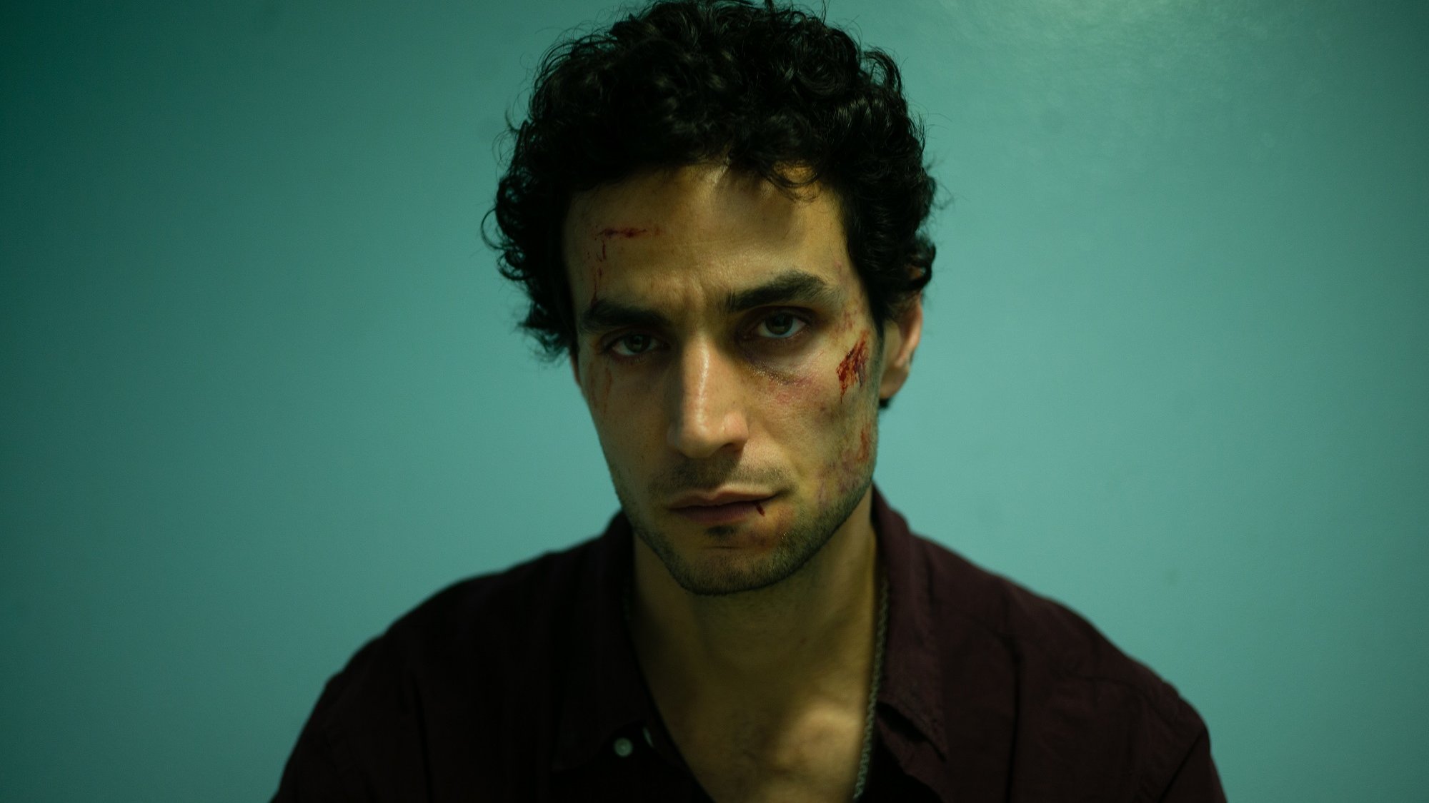 L'image montre un homme assis devant un mur de couleur bleu clair. Il a des cheveux noirs et bouclés, et son expression faciale semble intense ou préoccupée. Son visage présente des éraflures ou des blessures visibles, ce qui suggère qu'il a traversé une situation difficile. Il porte une chemise foncée et a l'air fatigué ou en détresse. L'éclairage est dramatique, mettant en avant ses traits et les blessures sur son visage.