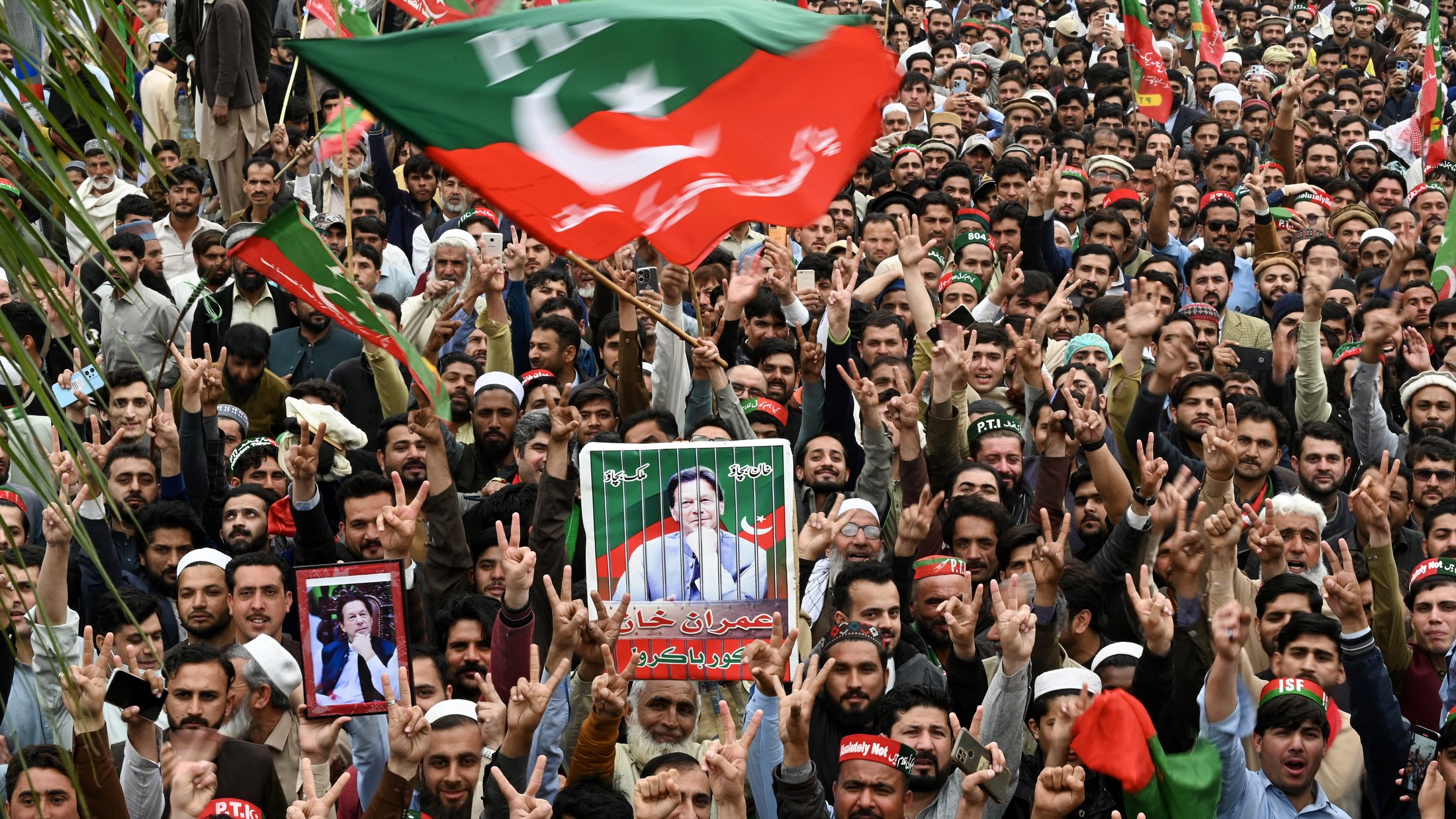 L'image montre une grande foule rassemblée lors d'un rassemblement. De nombreuses personnes brandissent des drapeaux, en particulier le drapeau du Parti Pakistan Tehreek-e-Insaf (PTI). On peut voir des manifestants levant les bras avec des gestes de victoire, ainsi que des pancartes et des portraits de leaders politiques. L'ambiance semble dynamique et engageante, avec des cris et des slogans de la part des partisans.