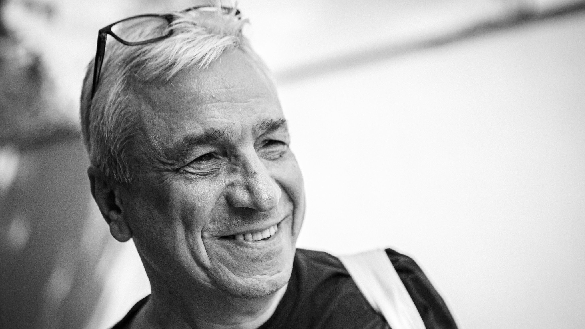 L'image montre un homme souriant. Il a des cheveux blancs et porte des lunettes sur le haut de sa tête. Son expression est joyeuse, et on peut sentir une atmosphère amicale. L'arrière-plan est flou, mettant davantage l'accent sur son visage. Il porte un t-shirt noir et semble détendu.