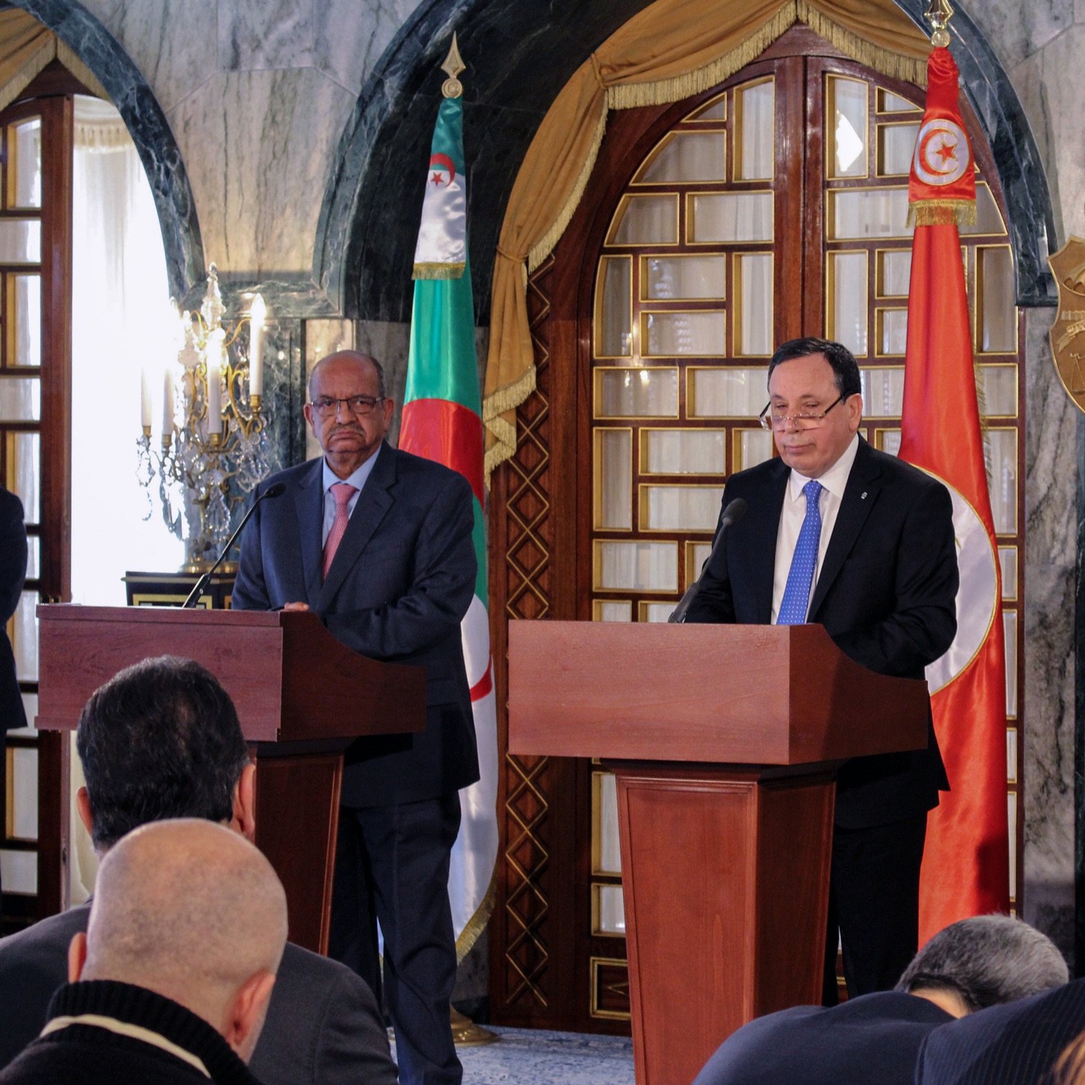 The image depicts a press conference or official meeting involving three individuals standing behind wooden podiums. The man in the center appears to be speaking, while the two flanking him are listening. The backdrop features flags of Algeria and Tunisia, along with a decorative emblem. The setting has an elegant, formal ambiance, likely within a government building. There are several journalists or attendees seated in front, capturing the event.