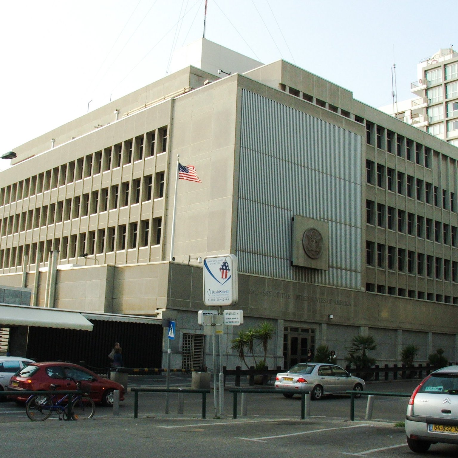 تظهر الصورة مبنى كبير متعدد الطوابق، يتميز بتصميمه المعماري العصري. المبنى يتكون من واجهات أسمنتية ونافذات كبيرة، ويظهر علم الولايات المتحدة الأمريكية مرفوعًا على أحد جوانبه. في الزاوية السفلية، توجد لافتة تشير إلى وجود مكان مخصص للسيارات، كما يظهر بعض الأشخاص والسيارات في الشارع المحيط. يبدو أن المبنى ينتمي إلى مؤسسة حكومية أو دبلوماسية.