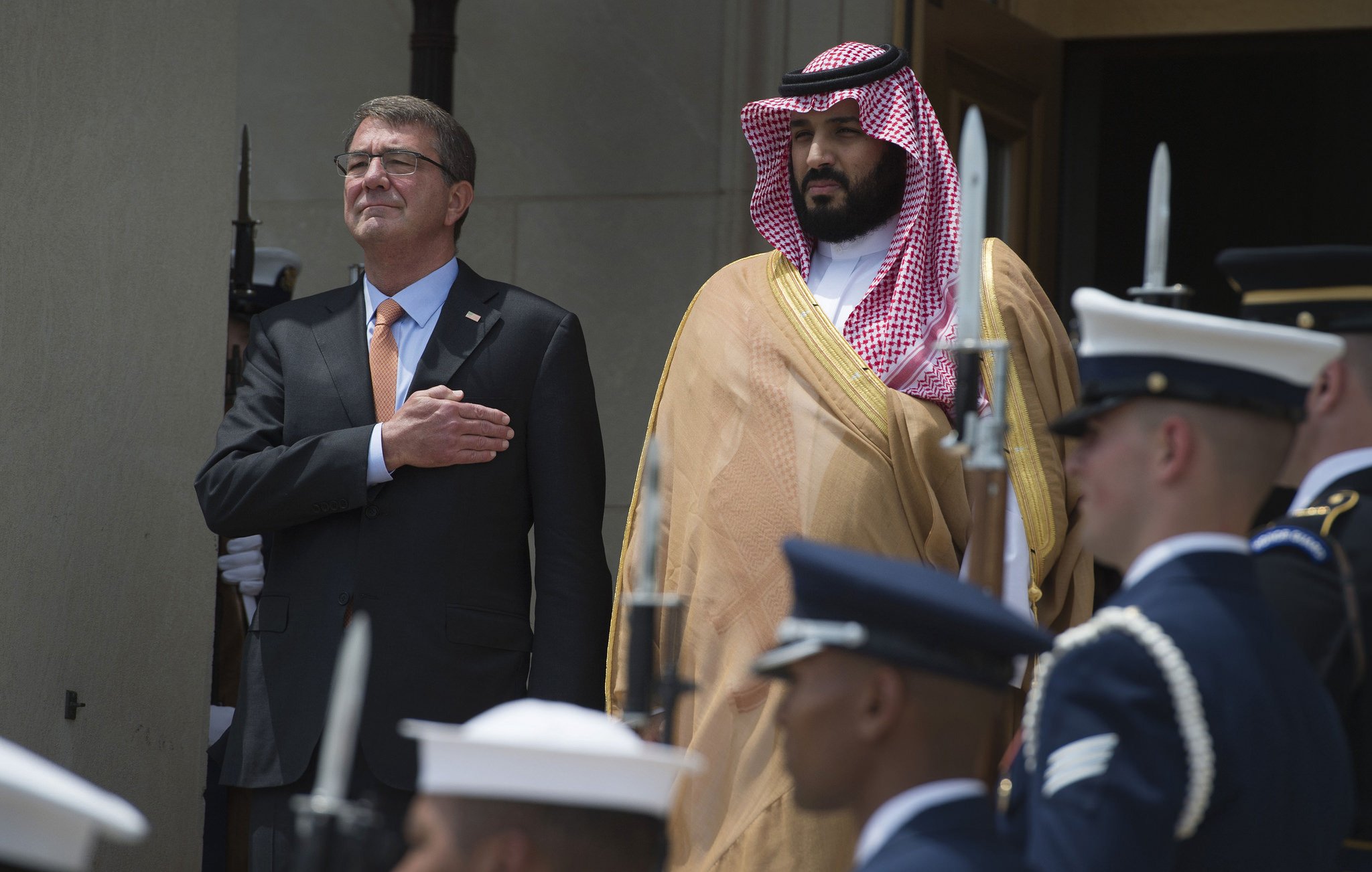 L'image montre deux hommes en tenue formelle. L'un est sur le devant, ayant la main sur le cœur, tandis que l'autre se tient à ses côtés. Ils sont entourés de membres d'une garde d'honneur, portant des uniformes militaires. L'environnement semble officiel, probablement un événement diplomatique ou militaire. Les deux hommes ont l'air sérieux et engagés dans la cérémonie.