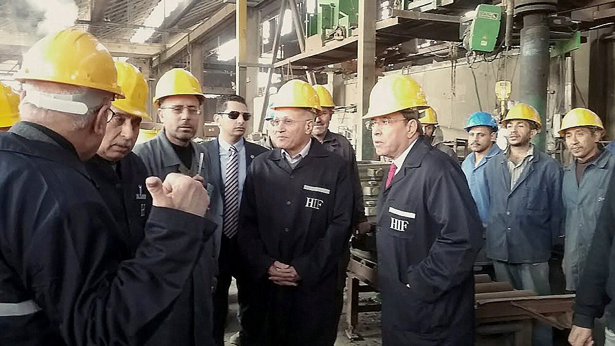 L'image montre un groupe de personnes dans un environnement industriel, probablement dans une usine. Les participants portent des casques de sécurité jaunes, ce qui indique qu'ils sont dans un lieu de travail où des précautions de sécurité sont nécessaires. Certains hommes sont en train de discuter ou d'écouter attentivement, tandis que d'autres observent la scène. En arrière-plan, on peut voir des machines et des équipements typiques d'un environnement de production. L'atmosphère semble être sérieuse et professionnelle.