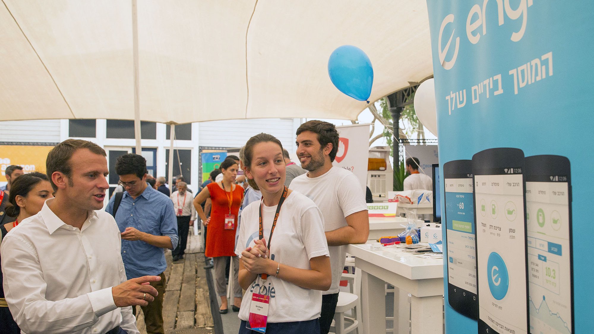 تظهر الصورة مشهداً في حدث أو معرض، حيث يتواجد عدد من الأشخاص تحت خيمة. يتمركز شخص يرتدي قميصاً أبيض، يتحدث مع شخص آخر يبدو أنه زائر. خلفهم، توجد لافتة تحمل شعار "engie" مع صور توضيحية لواجهة تطبيقات على الهواتف. يظهر في الصورة أيضاً عدد من الأشخاص الآخرين في الخلفية، وبعضهم يرتدي ملابس تحمل شارات. الجو يبدو غير رسمي ومليئاً بالنشاط.