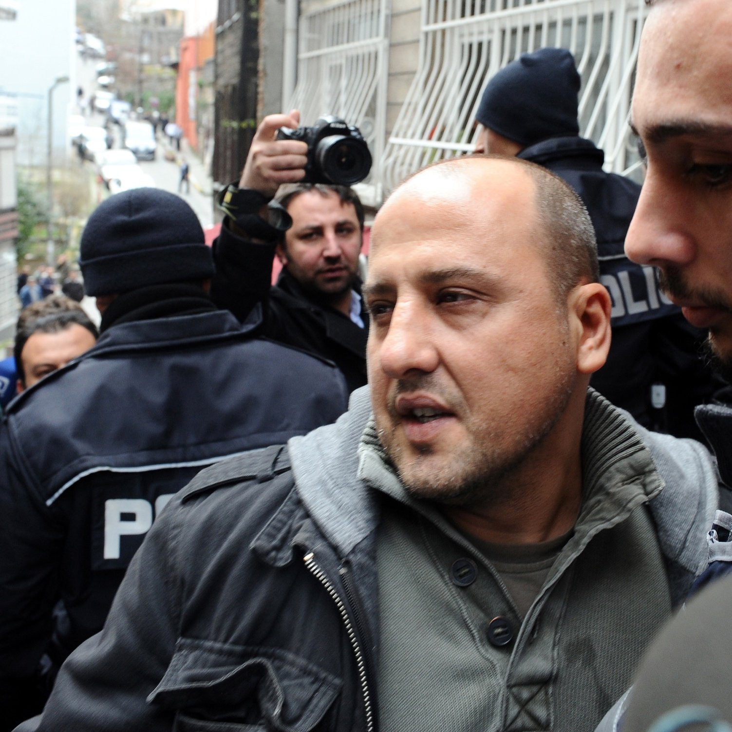 The image depicts a scene with a group of people, likely in a city setting. In the foreground, two men are present. One man, appearing distressed or concerned, is being escorted by another, who seems to be in a position of authority. The background includes police officers and a crowd, indicating a tense situation or an event such as a protest or arrest. The overall atmosphere appears to be one of heightened emotion or urgency.