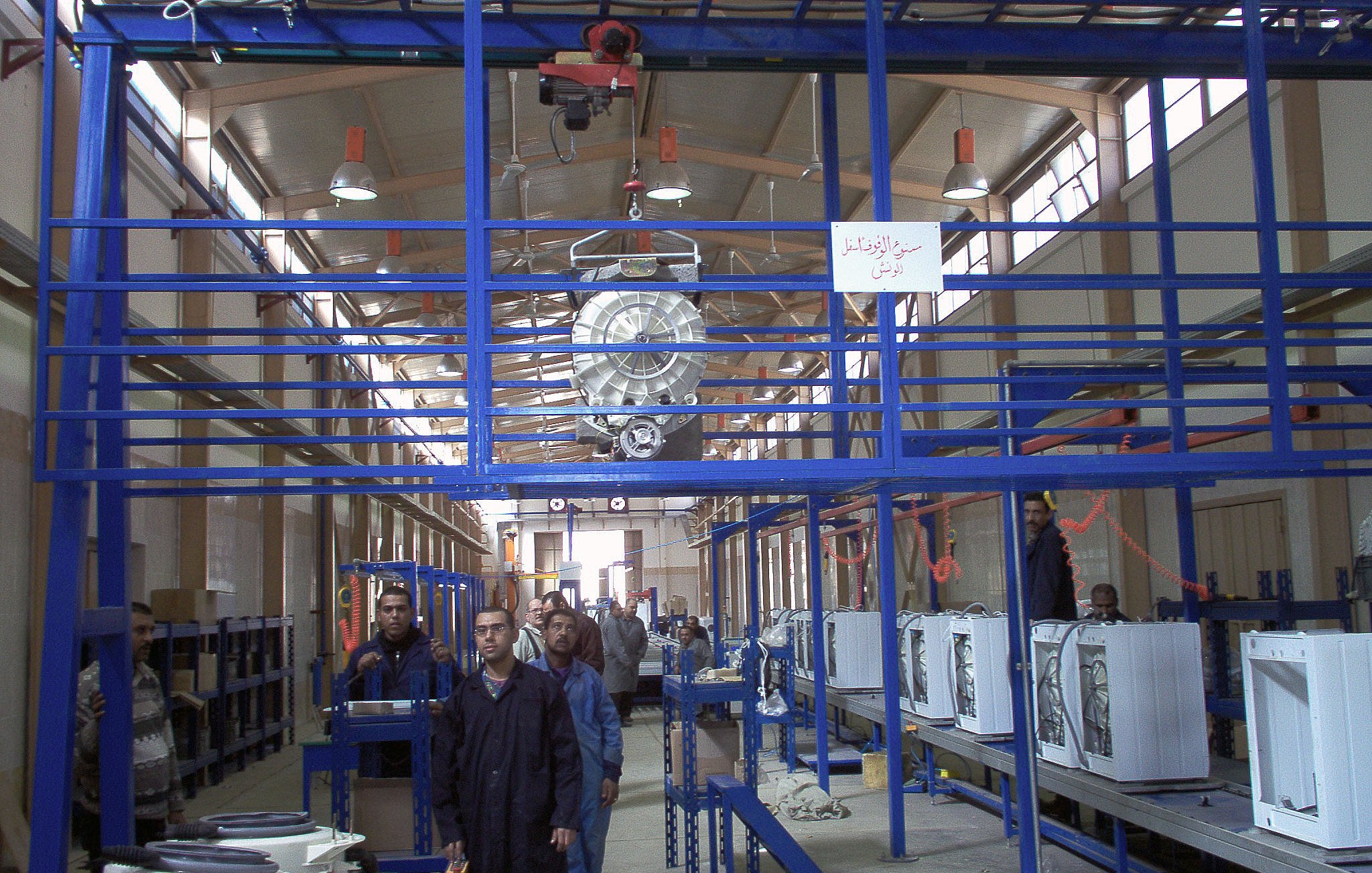 The image shows an industrial setting, likely a manufacturing or assembly facility. There are several workers in the foreground, wearing uniforms, and they seem to be engaged in tasks related to assembling or inspecting products. Above them, there is overhead equipment, possibly a crane or hoist, used for lifting materials. The facility has rows of workstations with what appears to be appliances or machinery on display, indicating a focus on production. The interior is well-lit with a spacious design, featuring high ceilings and a structured layout.