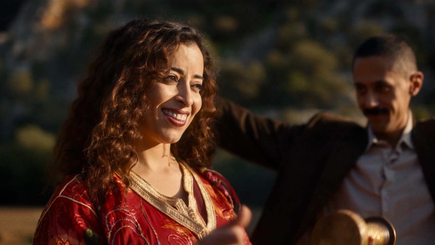 L'image montre une femme souriante avec des cheveux bouclés, portant une robe rouge ornée de motifs. Elle semble joyeuse et est entourée d'un environnement naturel. Un homme en arrière-plan, vêtu d'un costume, semble également profiter du moment. L'atmosphère générale est festive et conviviale.