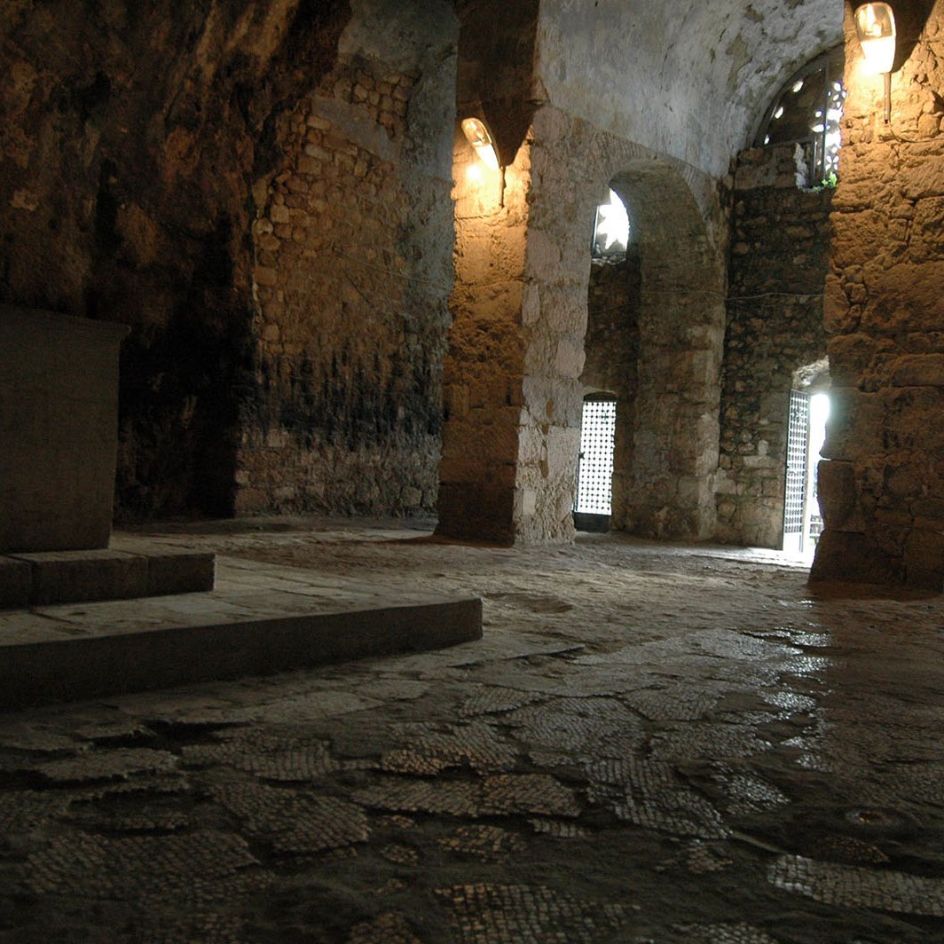 The image depicts an interior space that appears to be a stone chamber or crypt. The walls are made of rough stone, showcasing an ancient architectural style. Light filters in through arched windows, casting a soft glow on the uneven stone floor. There are several columns supporting the ceiling, and a platform or altar can be seen against one wall. The atmosphere is quiet and austere, suggesting a place of historical or spiritual significance.