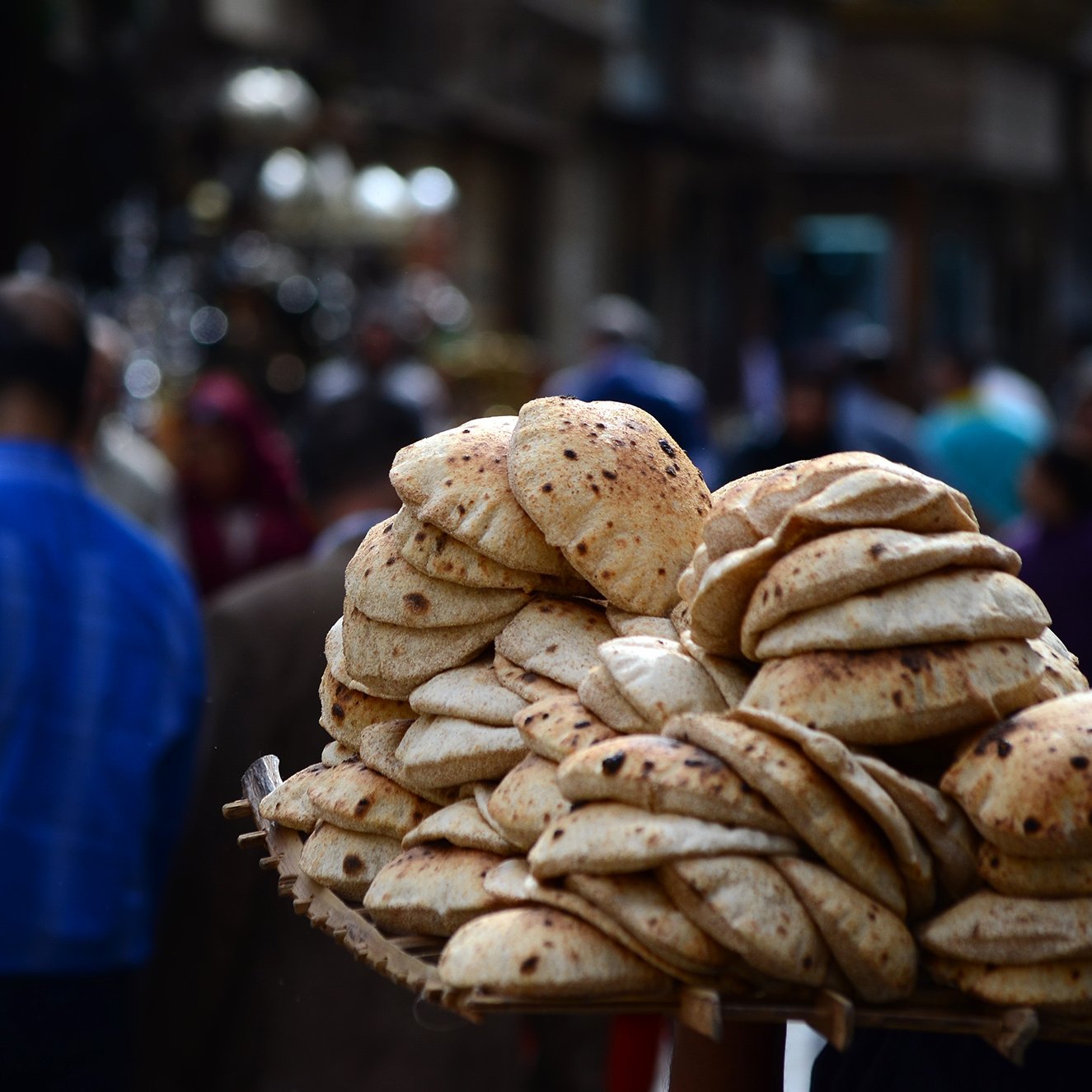 تظهر الصورة مجموعة من الخبز الطازج، يبدو أنه خبز عربي، مُتراص بشكل جميل في سلة يحملها شخص. في الخلفية، يتواجد عدد من الأشخاص في شارع مزدحم، مما يوحي بأجواء نابضة بالحياة في المدينة. يركز المشهد على الخبز كجزء من الثقافة الغذائية.