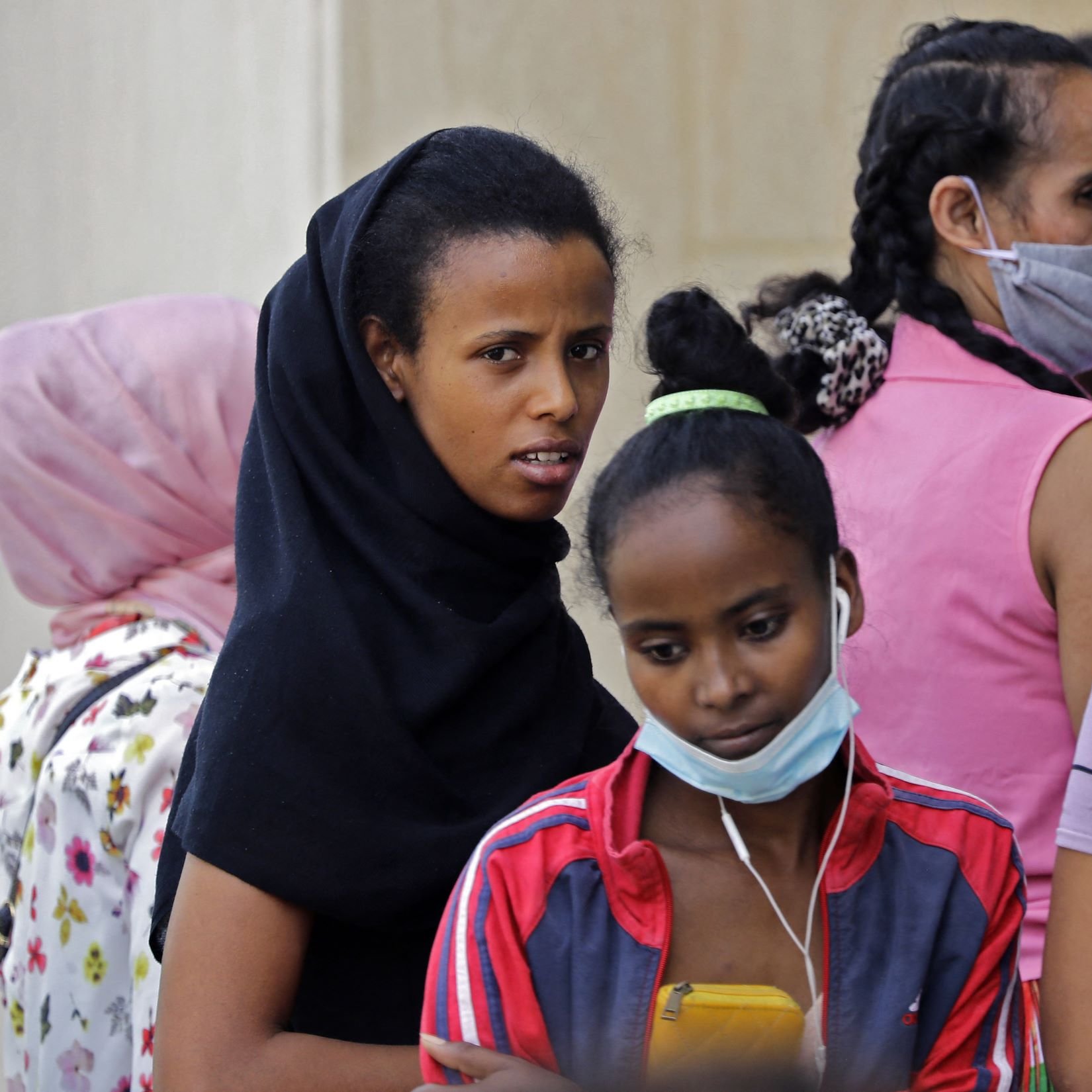 La imagen muestra a un grupo de personas, principalmente mujeres y niñas, en un entorno cercano. Algunas de ellas llevan mascarillas y visten ropa variada. Una de las mujeres en el primer plano lleva un velo negro y parece estar mirando hacia la cámara con una expresión seria. Otra niña, que se encuentra justo delante de ella, sostiene un teléfono móvil y lleva unos auriculares. El fondo incluye a más mujeres que están en la misma área, algunas de las cuales están de espaldas. La escena transmite un sentido de comunidad y concentración en la actividad que están realizando.