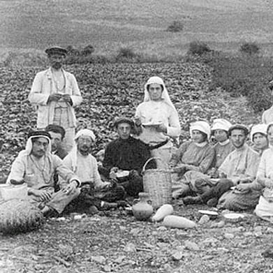 The image depicts a group of people gathered in an agricultural setting, likely in the early 20th century based on the clothing styles. There are both men and women, some wearing traditional garments and hats. They appear to be taking a break from farming activities, sitting on the ground with various tools and baskets nearby. The landscape in the background features fields and hills, suggesting a rural environment. Overall, it captures a moment of community among laborers.