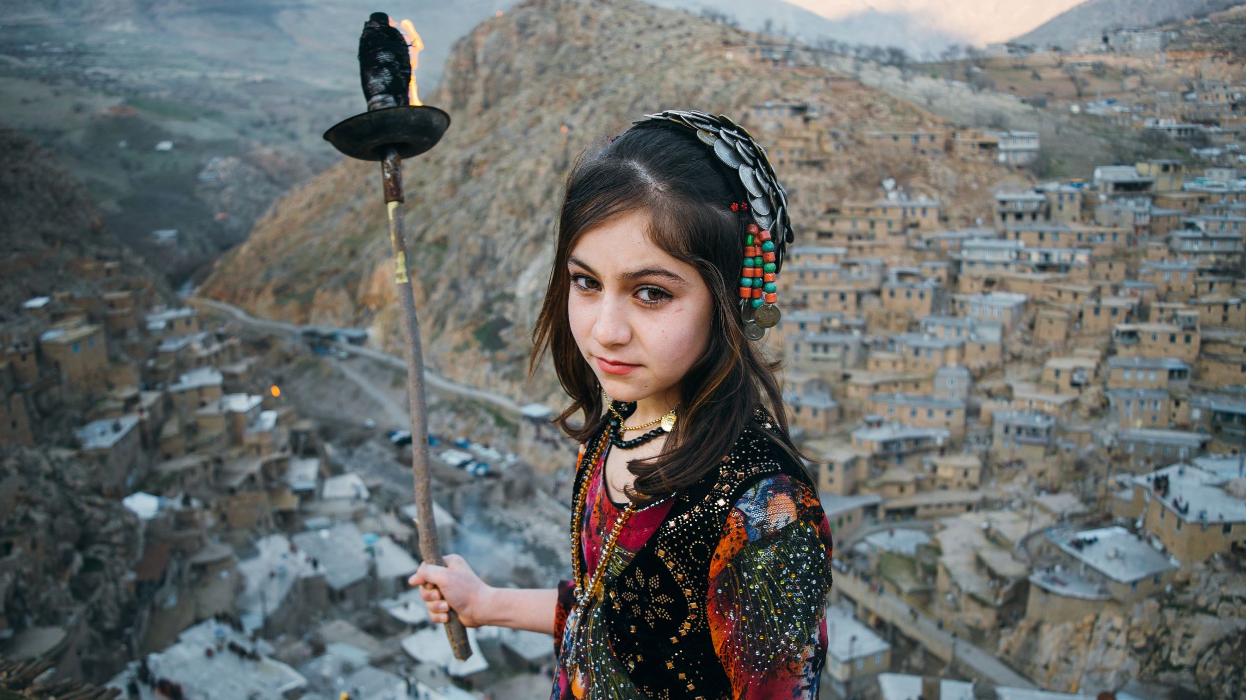 L'image montre une jeune fille tenant une torche, se tenant au sommet d'une colline surplombant un village pittoresque. Elle porte des vêtements colorés ornés de motifs traditionnels, et son regard est à la fois sérieux et perçant. En arrière-plan, on aperçoit des maisons construites à flanc de montagne, avec des paysages verdoyants s'étendant au loin. L'atmosphère de la scène est à la fois authentique et captivante, évoquant des traditions anciennes et une culture riche.