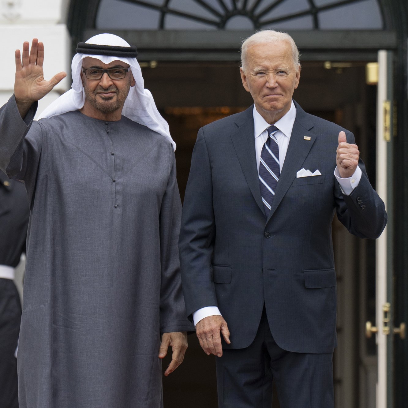L'image montre deux hommes se tenant à l'extérieur d'un bâtiment, probablement la Maison Blanche. L'un des hommes est vêtu d'une robe traditionnelle, un keffieh sur la tête, et fait un geste de la main, tandis que l'autre porte un costume avec une cravate. Derrière eux, des membres des forces armées en uniforme saluent. L'ambiance semble formelle, suggérant un événement diplomatique ou une rencontre officielle.