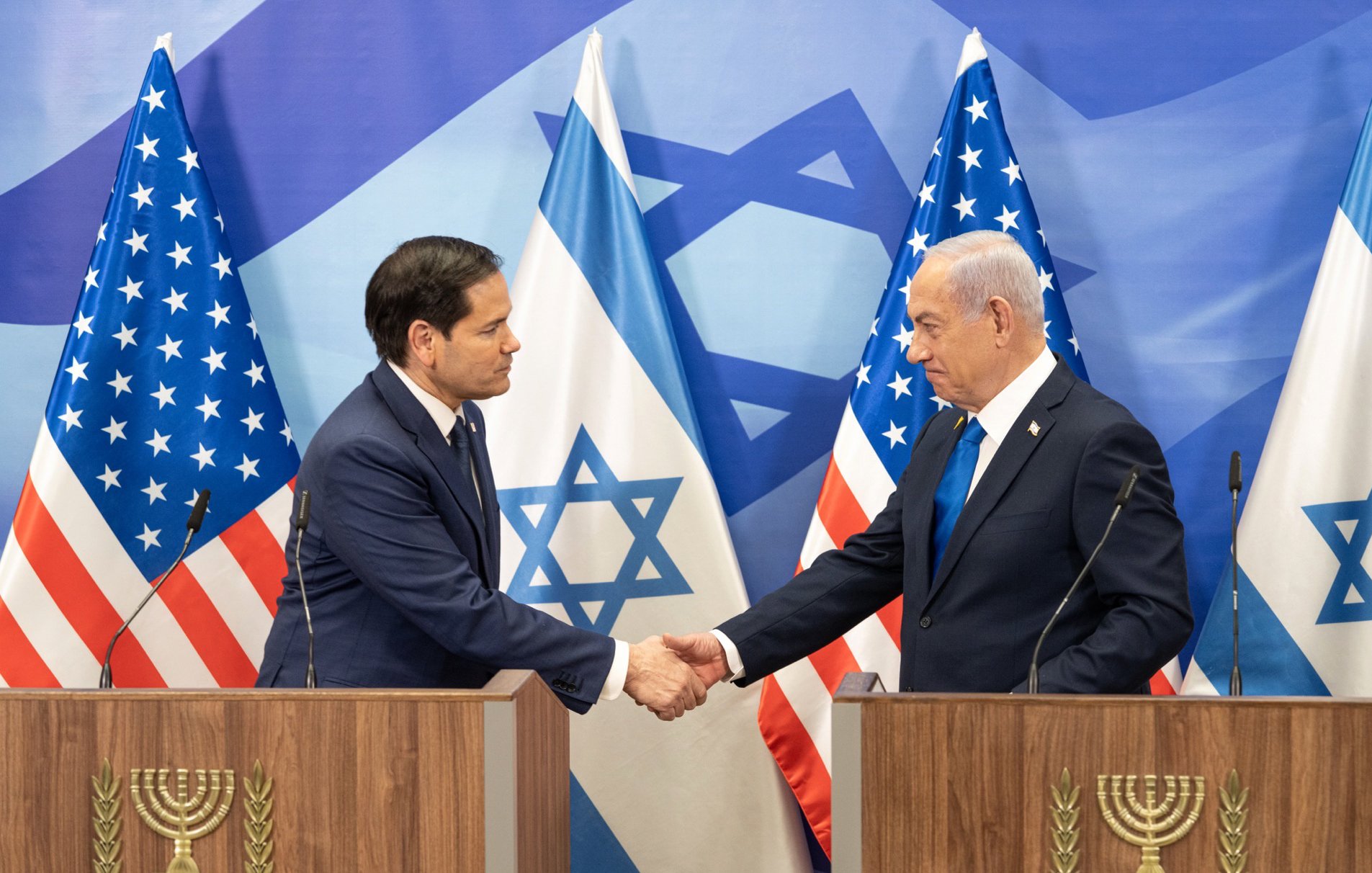 The image shows two men shaking hands at a press conference. They are standing at podiums, with Israeli and American flags in the background.