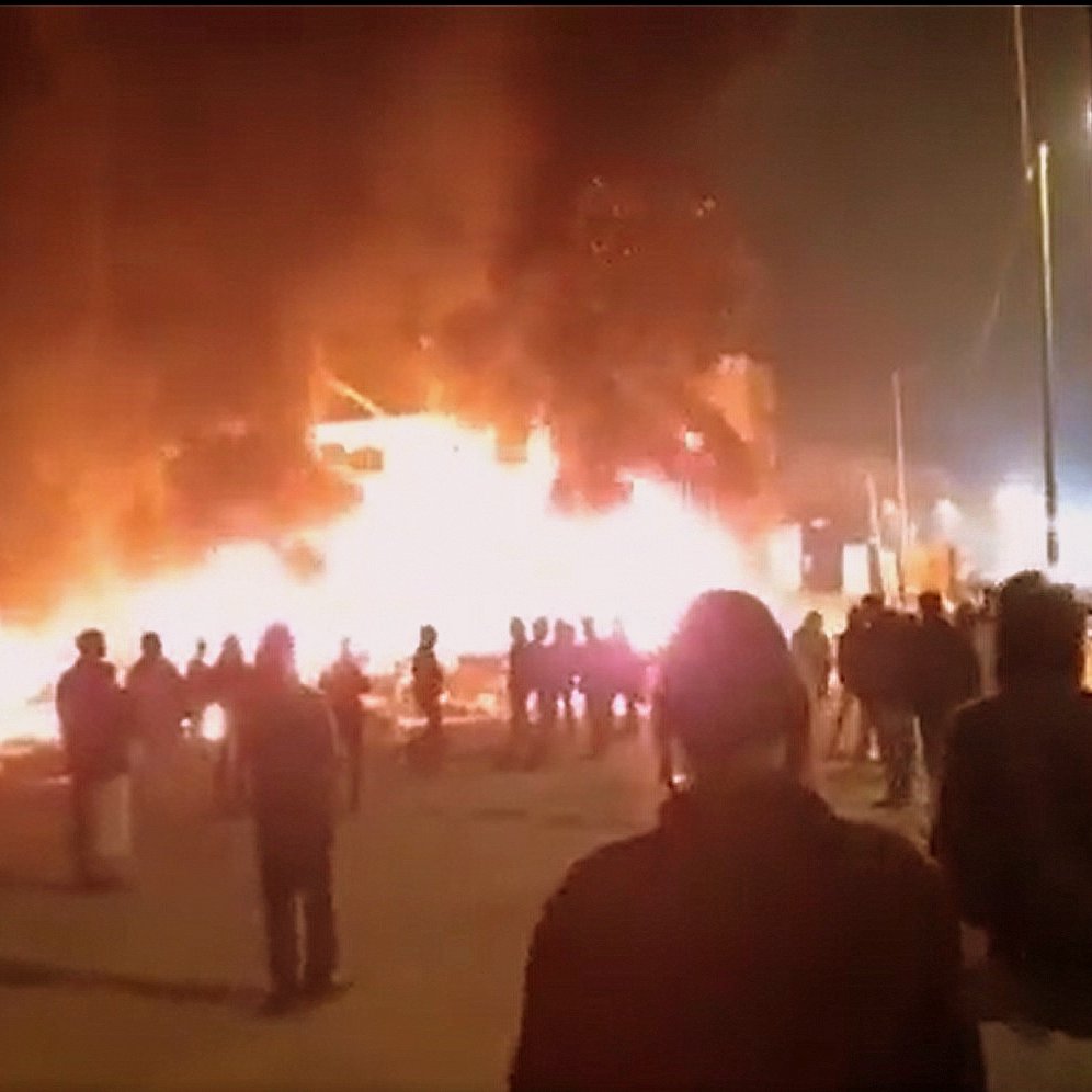 L'immagine mostra una scena notturna in cui un edificio è avvolto dalle fiamme, creando una grande quantità di fumo e luce. Davanti all'edificio in fiamme, un gruppo di persone si raduna, osservando la situazione. Le figure sembrano essere in lontananza, mentre il fuoco illumina l'ambiente circostante. È possibile notare un'atmosfera di tensione e allerta.