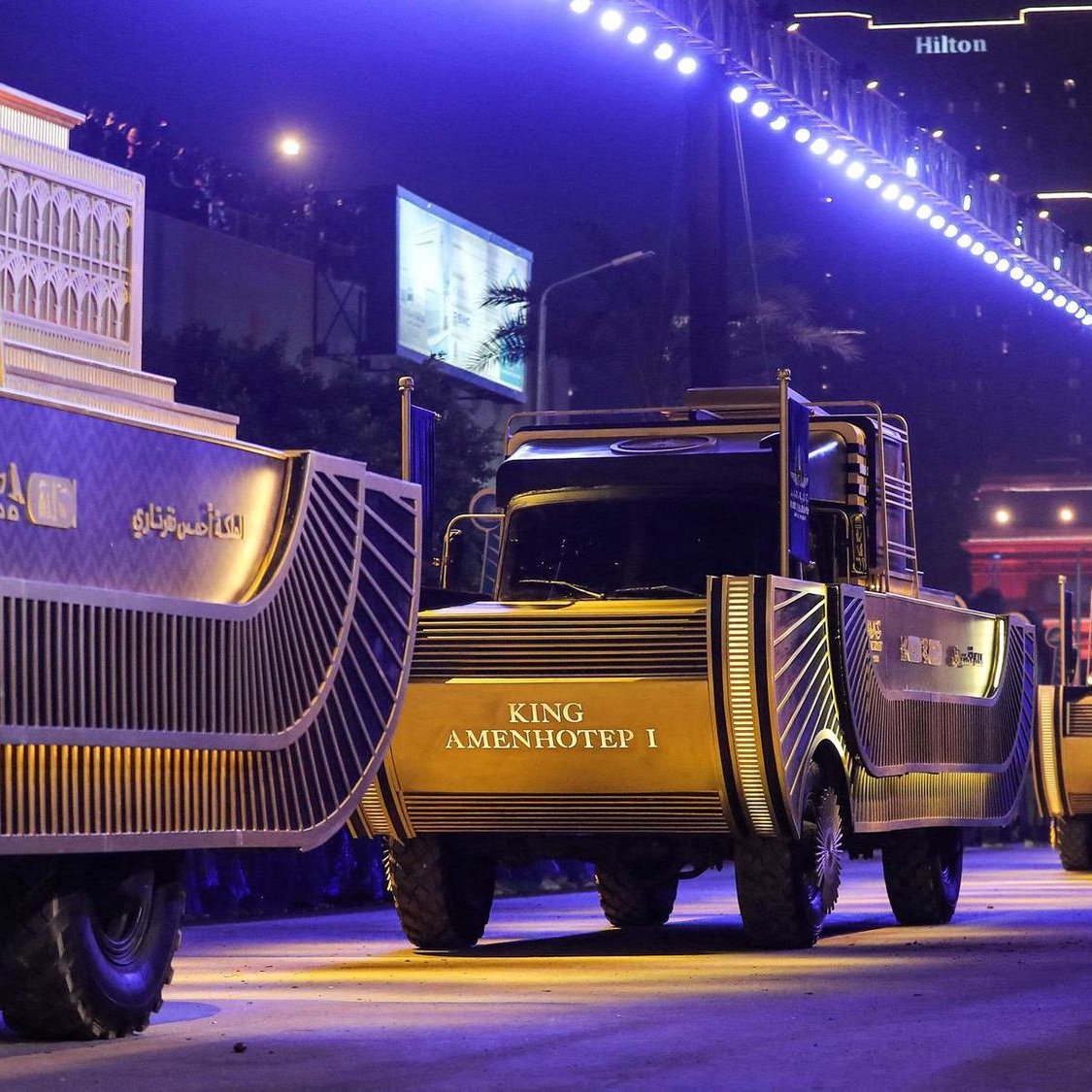 La imagen muestra una escena nocturna de un desfile en la que varios camiones decorados están alineados en una calle iluminada. Los camiones tienen un diseño llamativo y parecen estar relacionados con temas históricos, específicamente sobre reyes egipcios, como "KING AMENHOTEP I". En el primer plano, hay una persona vestida con ropa tradicional que sostiene un objeto, posiblemente una ofrenda o un instrumento musical. El ambiente es festivo y majestuoso, con luces brillantes que realzan el ambiente del evento. En el fondo, se pueden observar edificios y carteles, lo que sugiere que el desfile está teniendo lugar en una ciudad moderna.