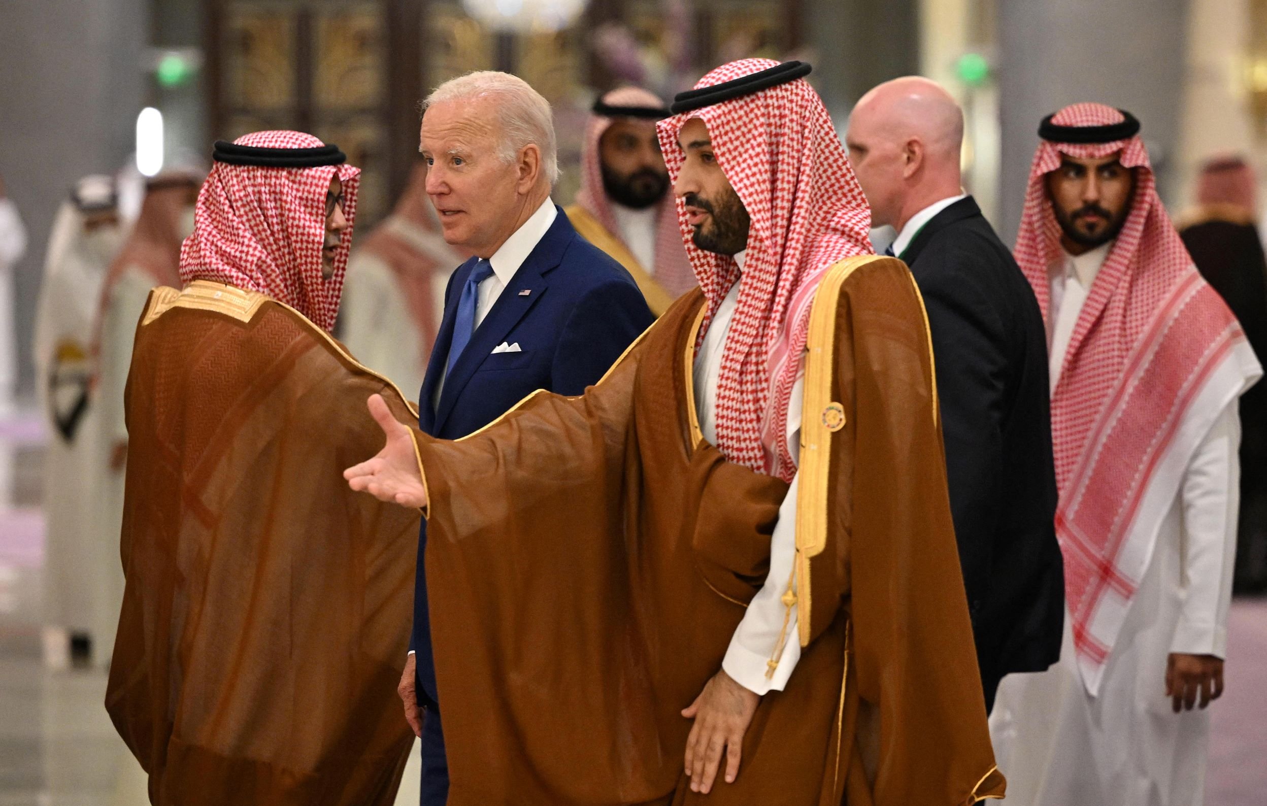 الصورة تظهر مجموعة من الأشخاص في مؤتمر أو حدث رسمي. في المقدمة، يوجد شخص يرتدي بدلة زرقاء، يبدو أنه يتحدث مع آخرين يرتدون الملابس التقليدية العربية. يتميز الأشخاص في الخلف بملابسهم الرسمية، ويتواجدون في بيئة داخلية أنيقة. التفاصيل تشير إلى أجواء من المفاوضات أو اللقاءات الدبلوماسية.