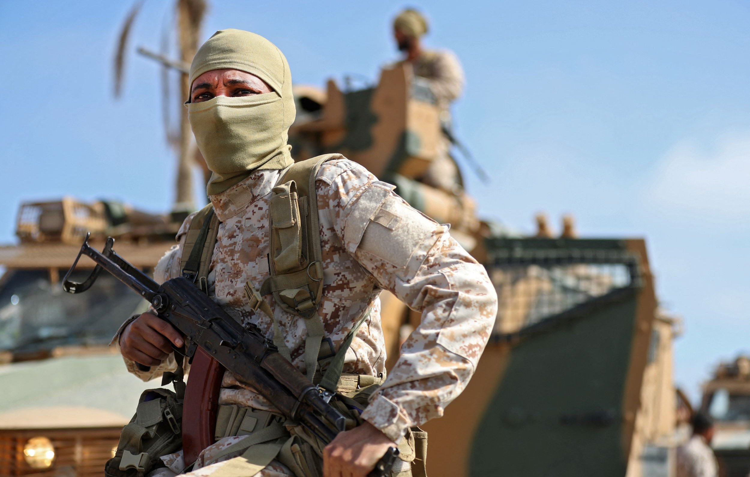 در این تصویر، یک سرباز مسلح با پوشش صورت، در حال ایستادن در پیش‌زمینه قرار دارد. او یونیفورم نظامی و کلاهی به سر دارد و یک اسلحه در دستش است. در پس‌زمینه، یک خودروی نظامی دیده می‌شود و تعدادی از سربازان دیگر در حال فعالیت هستند. آسمان آبی و آب و هوای روشن، صحنه را روشن می‌کند. این تصویر حس قدرت و دقت نظامی را به تصویر می‌کشد.