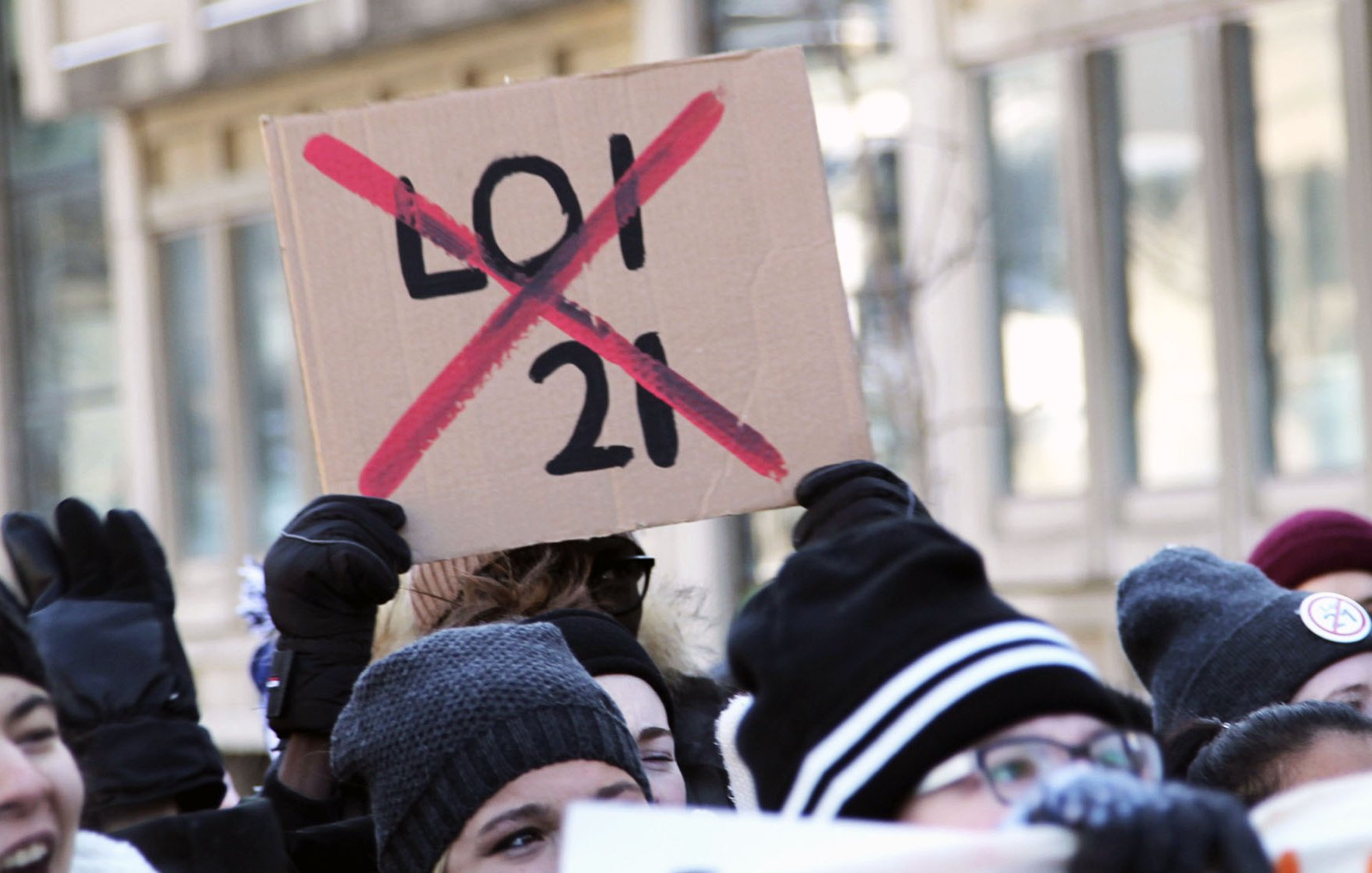 The image depicts a group of people participating in a protest. One prominent sign held up by a protester is made of cardboard and has "LOI 21" (with a red "X" crossing it out) written in bold letters. The crowd appears to be wearing winter clothing, such as hats and gloves, indicating that the protest is taking place in a cold environment. The atmosphere seems to be one of solidarity and activism, as the participants are visibly engaged in the demonstration.