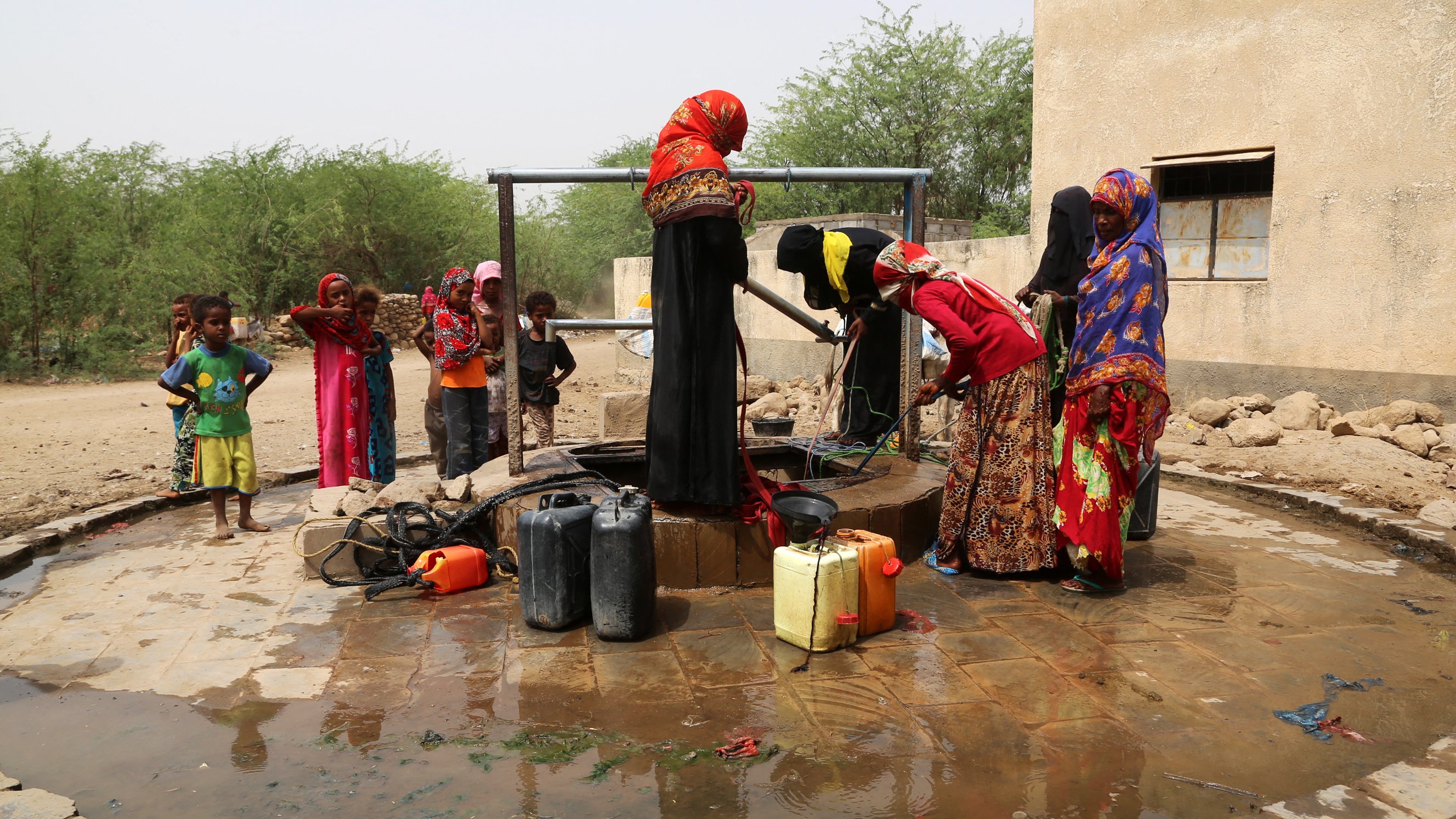 L'image montre un groupe de femmes près d'un puits. Elles sont vêtues de robes colorées et semblent être en train de remplir des conteneurs d'eau. Des enfants jouent autour d'elles. Le sol est humide, et le cadre semble rural, avec des buissons et une structure en arrière-plan. L'atmosphère semble active et communautaire, illustrant l'importance de l'accès à l'eau dans la vie quotidienne.
