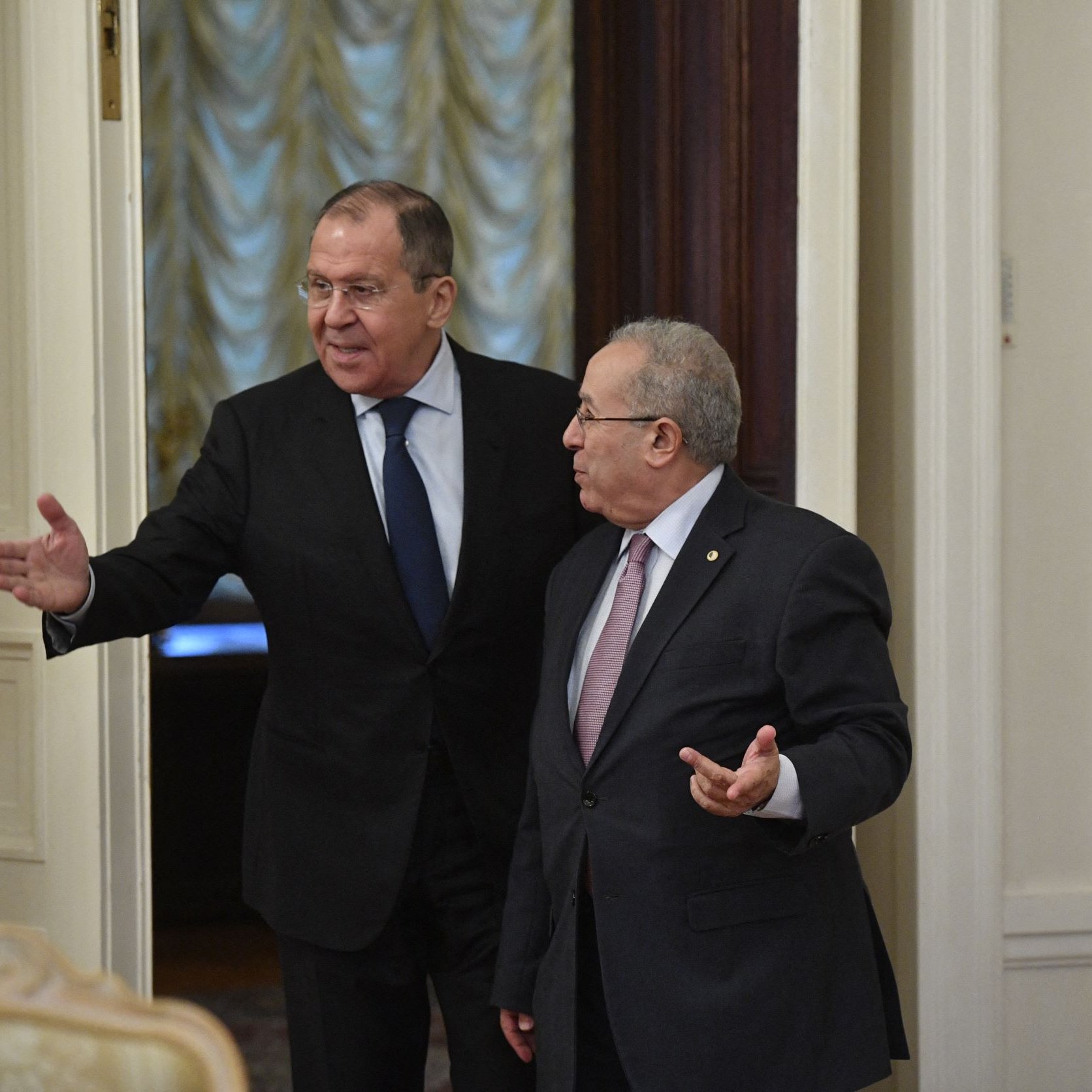 En la imagen se puede ver a dos hombres en un entorno formal, probablemente en una sala de reuniones o un despacho. Uno de ellos, a la izquierda, parece estar señalando o gesticulando con una mano, mientras que el otro, a la derecha, escucha y gira ligeramente su cuerpo hacia él. Ambos visten trajes formales y tienen expresiones serias. El ambiente parece elegante, con detalles en las paredes y una iluminación suave.