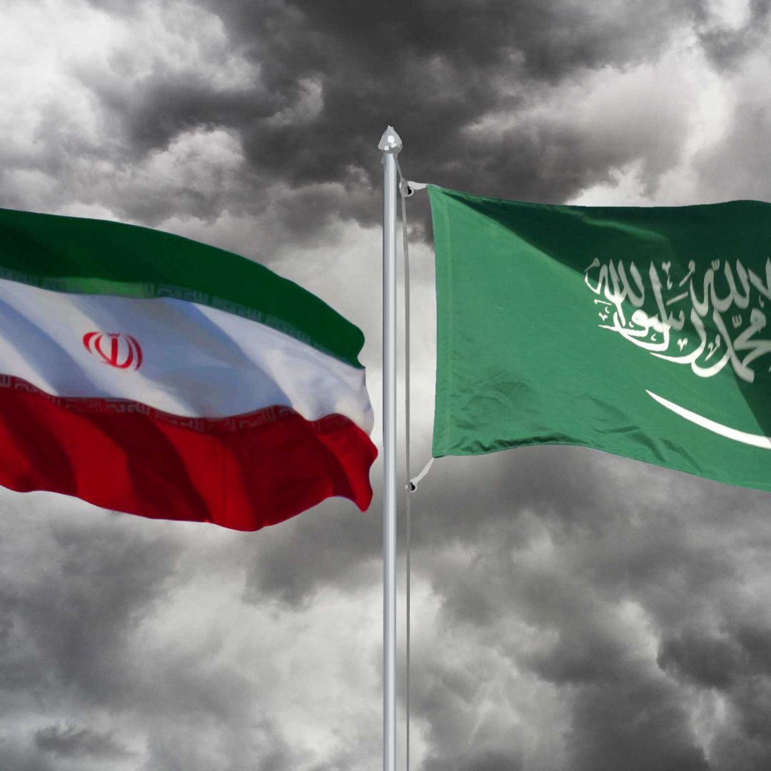 L'image montre deux drapeaux flottant l'un à côté de l'autre. À gauche, le drapeau iranien, qui est composé de bandes horizontales rouges, blanches et vertes, ainsi qu'un insigne en vert. À droite, le drapeau saoudien, qui est principalement vert avec un texte en calligraphie arabe en blanc et une épée en dessous. En arrière-plan, on peut voir des nuages sombres, ce qui crée une ambiance dramatique.