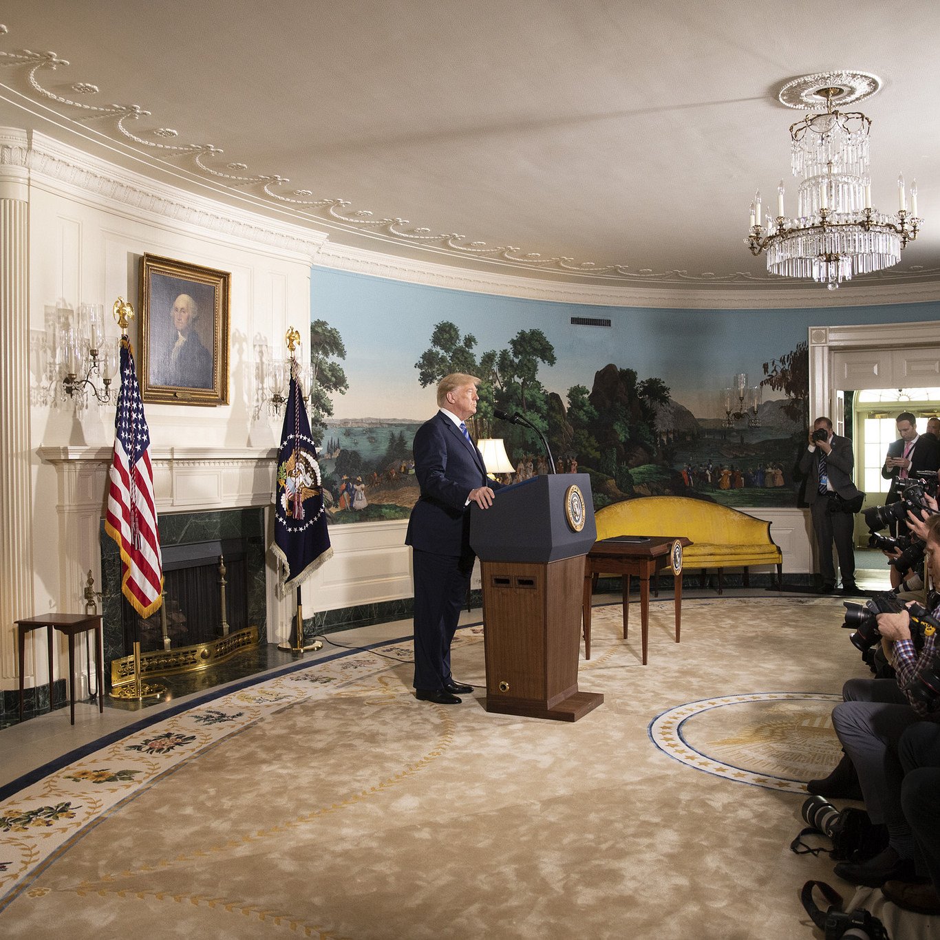 L'image montre un homme debout devant un podium, probablement lors d'une allocution publique. Il est situé dans une pièce décorée avec des murs peints de paysages, et des drapeaux américain et présidentiel sont visibles à côté de lui. Plusieurs journalistes et photographes sont présents, capturant l'événement. Le mobilier de la pièce semble ancien, et il y a un grand lustre au plafond.