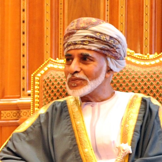 The image features a man seated in a richly decorated chair, wearing traditional attire. He is dressed in a white dishdasha, complemented by a decorative cape, and a turban, which is traditional in some Middle Eastern cultures. The background consists of intricate woodwork and ornate design elements, suggesting an opulent setting. The man's expression appears calm and thoughtful.