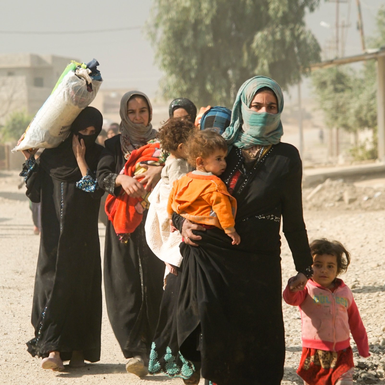 در این تصویر، گروهی از زنان و کودکان در یک منطقه روستایی دیده می‌شوند. زنان با لباس‌های سنتی و چادر، برخی از آنها با کودکان در آغوش و دیگران با بارهایی بر دوش در حال حرکت هستند. در پس‌زمینه، چند مرد هم دیده می‌شوند که به سمت دورتر در حال رفتن هستند. فضای تصویر خاکی و کم‌نور به نظر می‌رسد که نشان‌دهنده شرایط احتمالی نامساعد است.