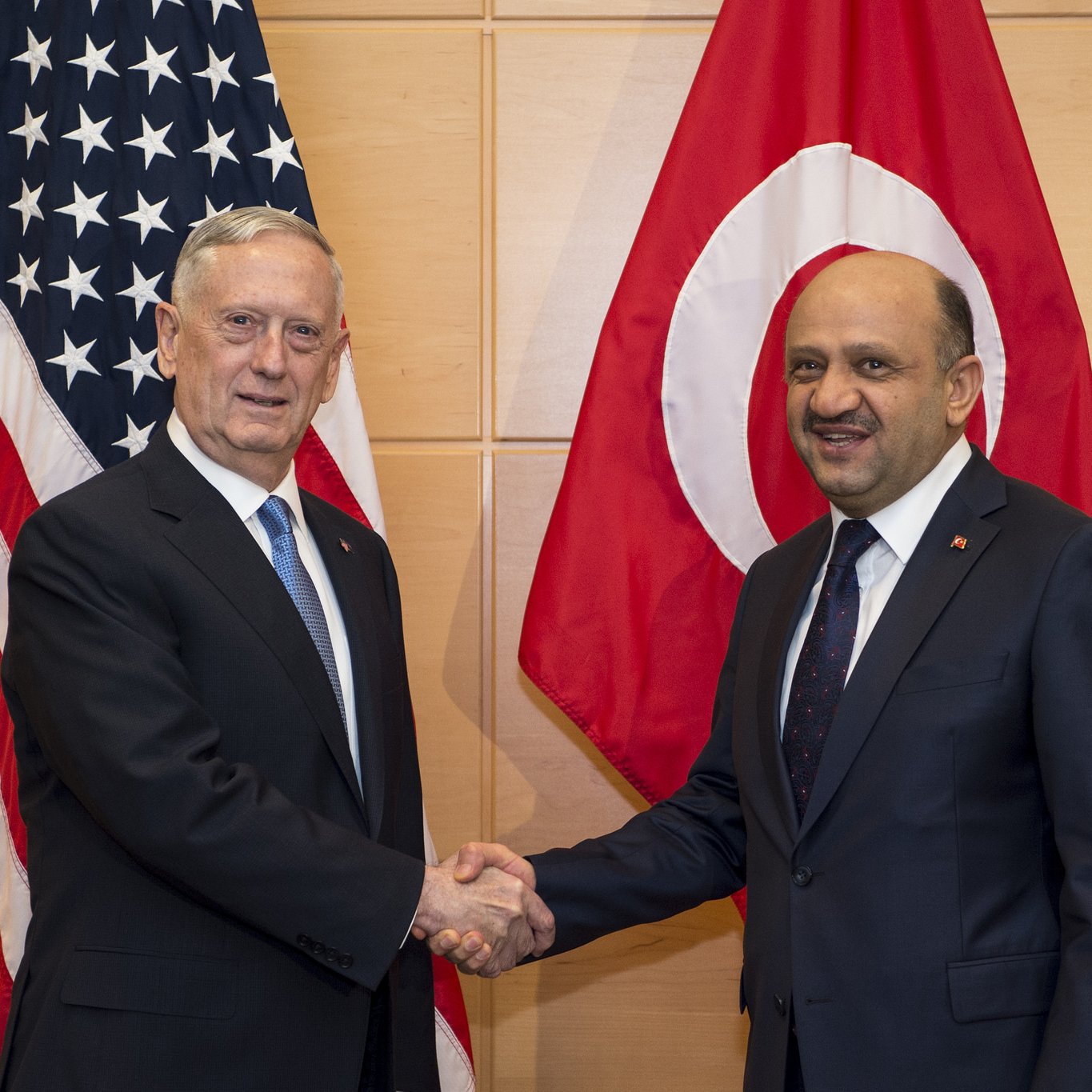 في الصورة، يظهر رجلان يتصافحان في مشهد رسمي. على اليمين، يوجد علم تركيا، وعلى اليسار يوجد علم الولايات المتحدة. يبدو أن الرجلين يتبادلان التحية، مما يشير إلى وجود تعاون أو لقاء دبلوماسي بين بلديهما.