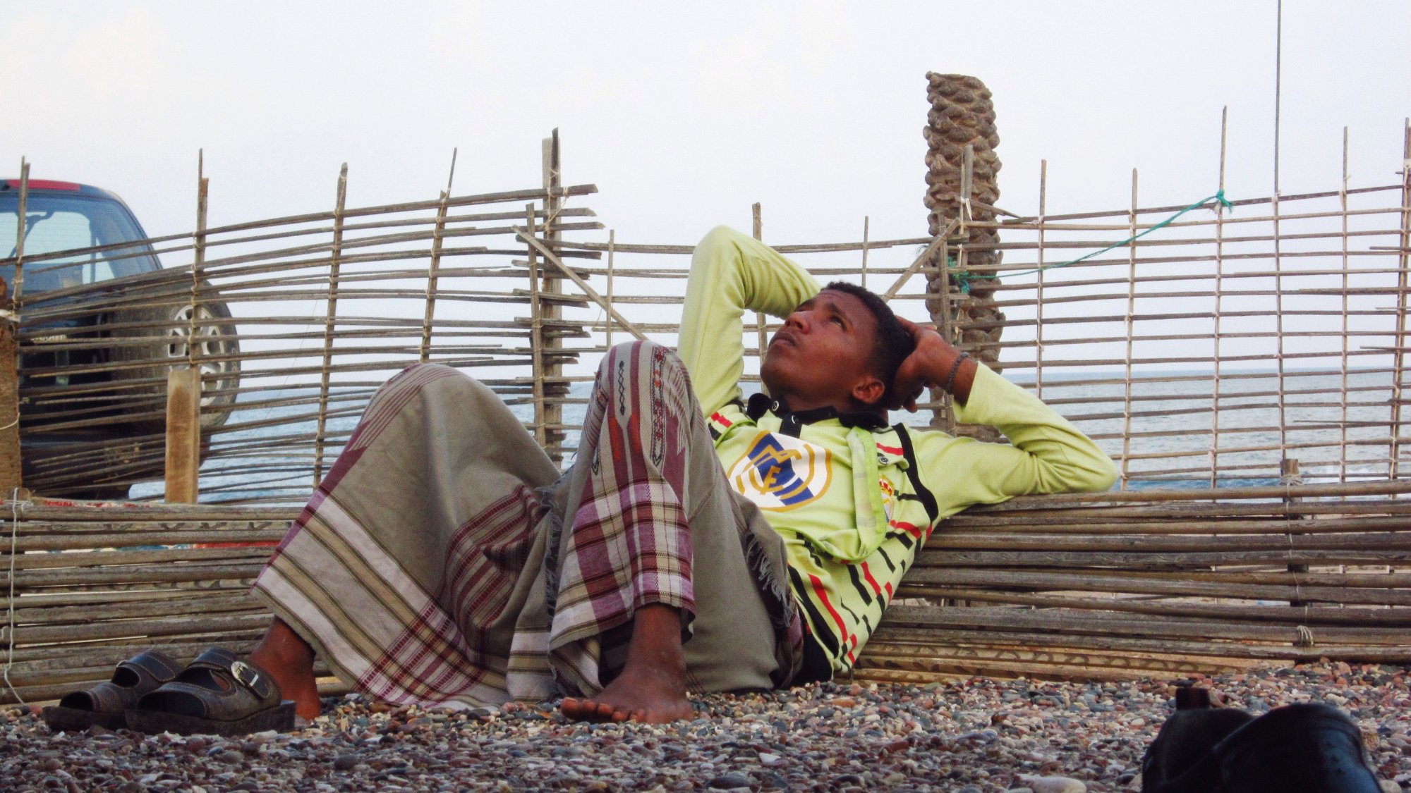 Un garçon allongé, pensif, contre une barrière en bois, sur une plage de gravier.