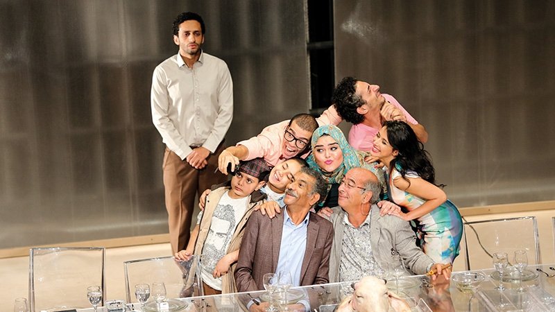 L'image montre un groupe de personnes rassemblées autour d'une table, apparemment lors d'un événement convivial. Il y a des adultes et des enfants, certains prenant la pose pour un selfie. Les expressions sont joyeuses et décontractées, avec des sourires sur presque tous les visages. À l'arrière-plan, un homme se tient debout, regardant la scène. La table est décorée avec des verres et un plat, ajoutant une ambiance festive à la scène. L'ensemble dégage une atmosphère d'amitié et de bonheur partagé.