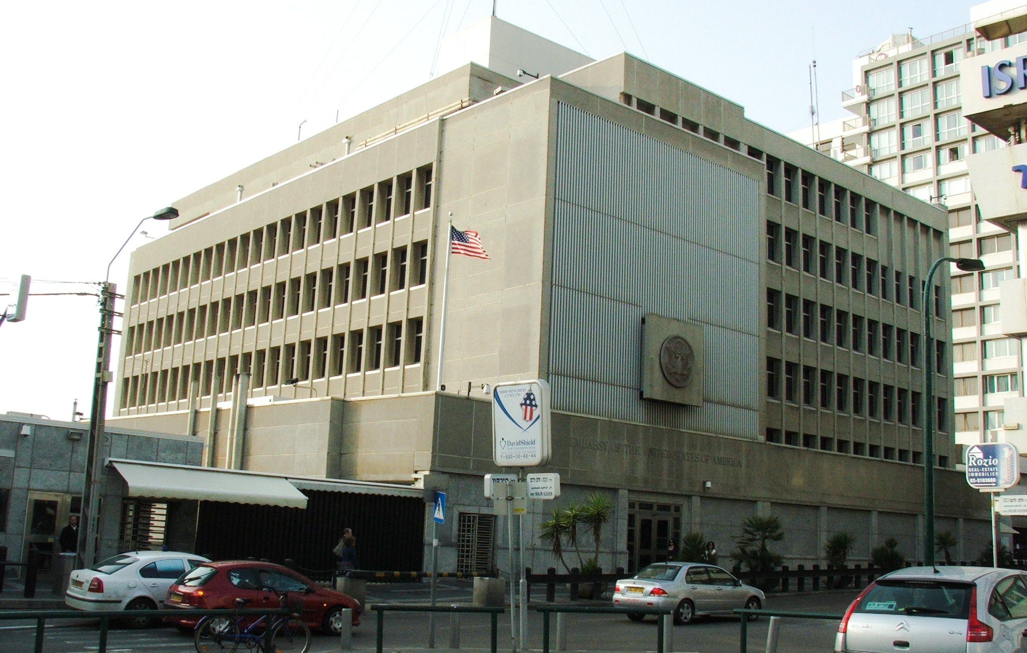 تظهر الصورة مبنى كبير متعدد الطوابق، يتميز بتصميمه المعماري العصري. المبنى يتكون من واجهات أسمنتية ونافذات كبيرة، ويظهر علم الولايات المتحدة الأمريكية مرفوعًا على أحد جوانبه. في الزاوية السفلية، توجد لافتة تشير إلى وجود مكان مخصص للسيارات، كما يظهر بعض الأشخاص والسيارات في الشارع المحيط. يبدو أن المبنى ينتمي إلى مؤسسة حكومية أو دبلوماسية.