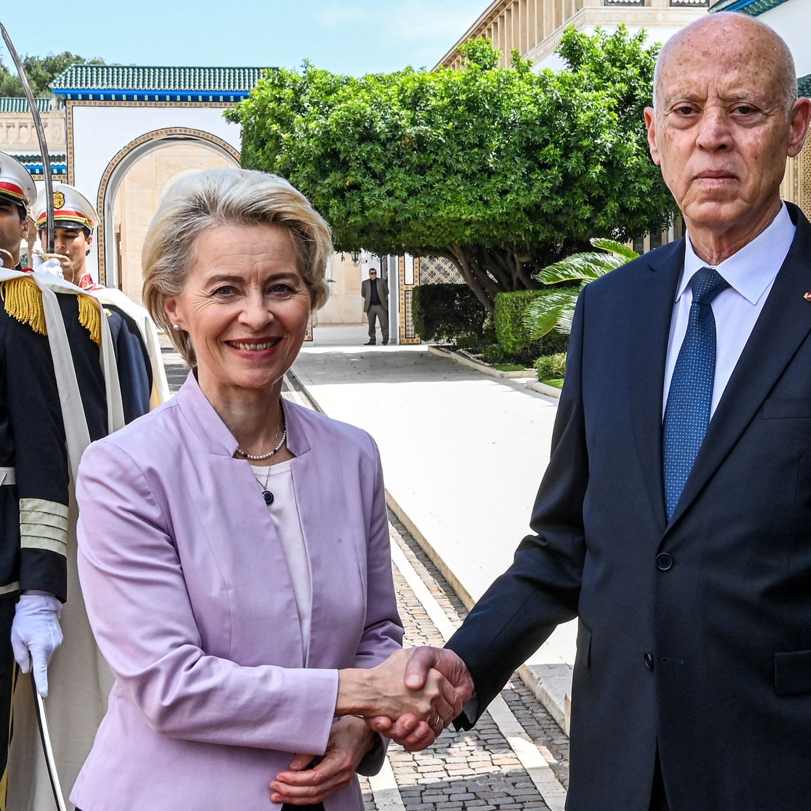 L'immagine mostra un incontro ufficiale tra due persone. Una donna, vestita con un abito chiaro, sorride mentre stringe la mano a un uomo in giacca e cravatta scura. Sullo sfondo, si possono vedere alcuni soldati in uniforme, che sembrano fare la guardia. La scena si svolge in un ambiente elegante, con piante e architetture raffinate visibili.