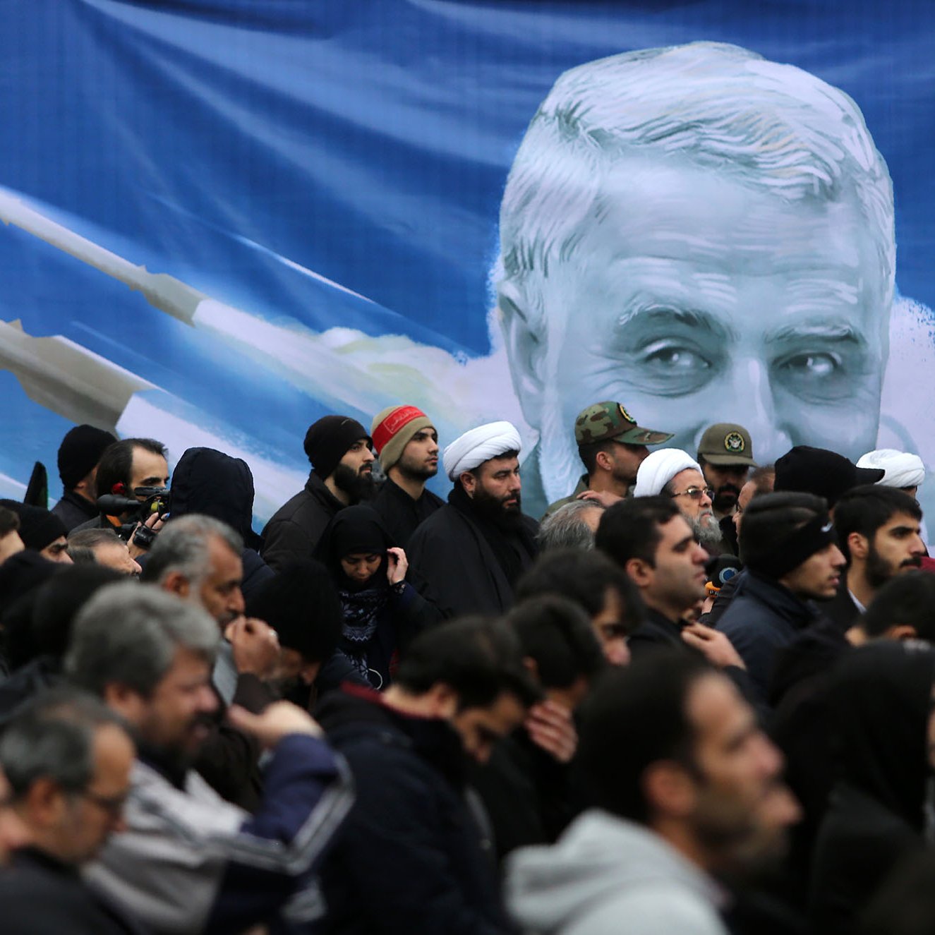 تصویر نشان‌دهنده تجمعی از افراد در یک مراسم است. مردم بیشتر لباس‌های تیره پوشیده‌اند و برخی از آن‌ها در حال گوش دادن به سخنان یا تماشای یک برنامه هستند. در پس‌زمینه، تصویری بزرگ از چهره‌ای که شبیه یک شخصیت شناخته‌شده است وجود دارد و در اطراف آن تصاویری از موشک‌ها دیده می‌شود. این تصویر به نوعی نمادین به نظر می‌رسد و می‌تواند به مناسبت‌های ملی یا مذهبی مربوط باشد. 