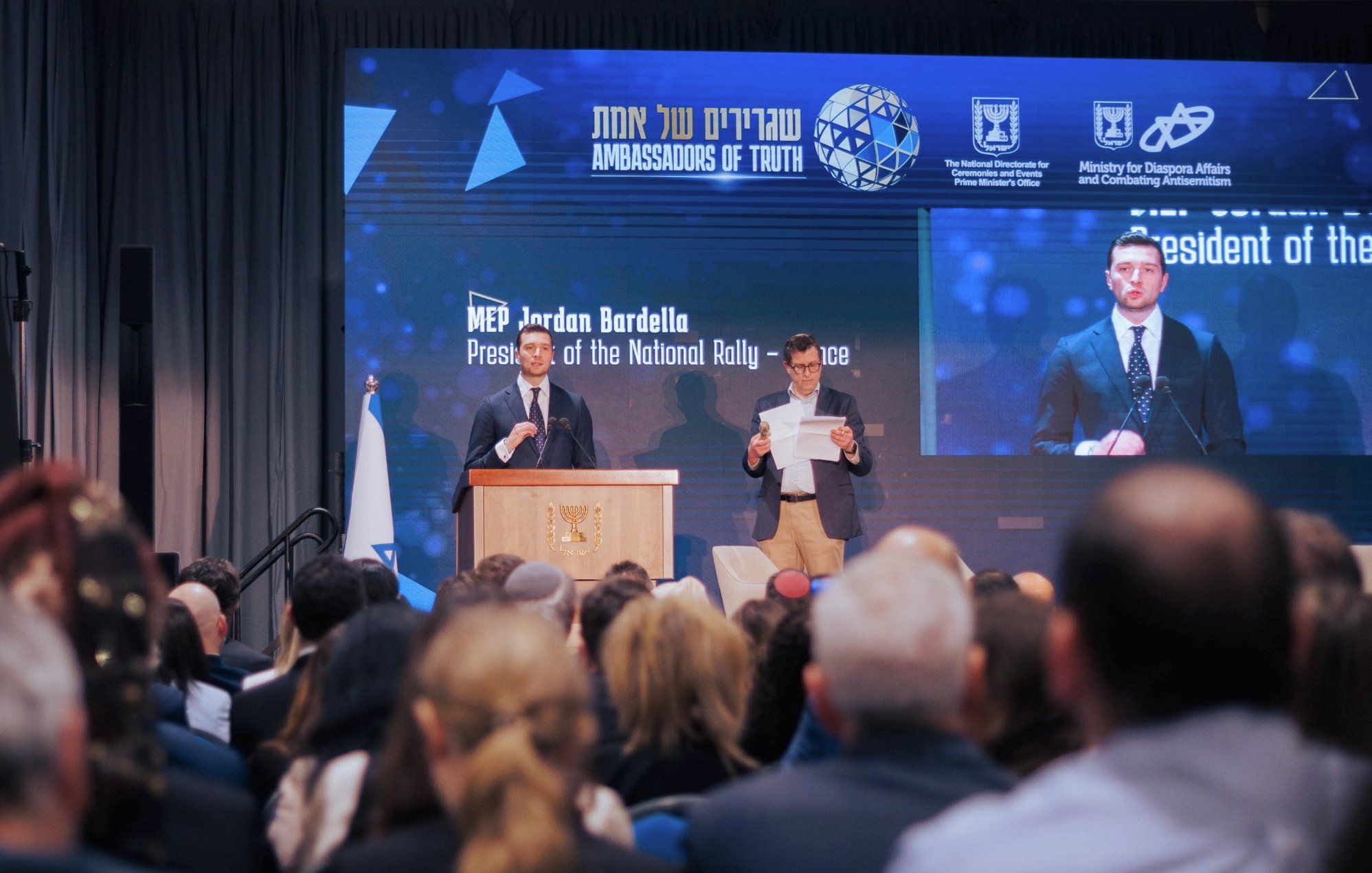 L'image montre une conférence avec un orateur debout derrière un pupitre, lisant un discours. Le public clairement visible est assis devant lui, et un grand écran derrière affiche des informations sur l'événement. L'atmosphère est formelle et engageante.