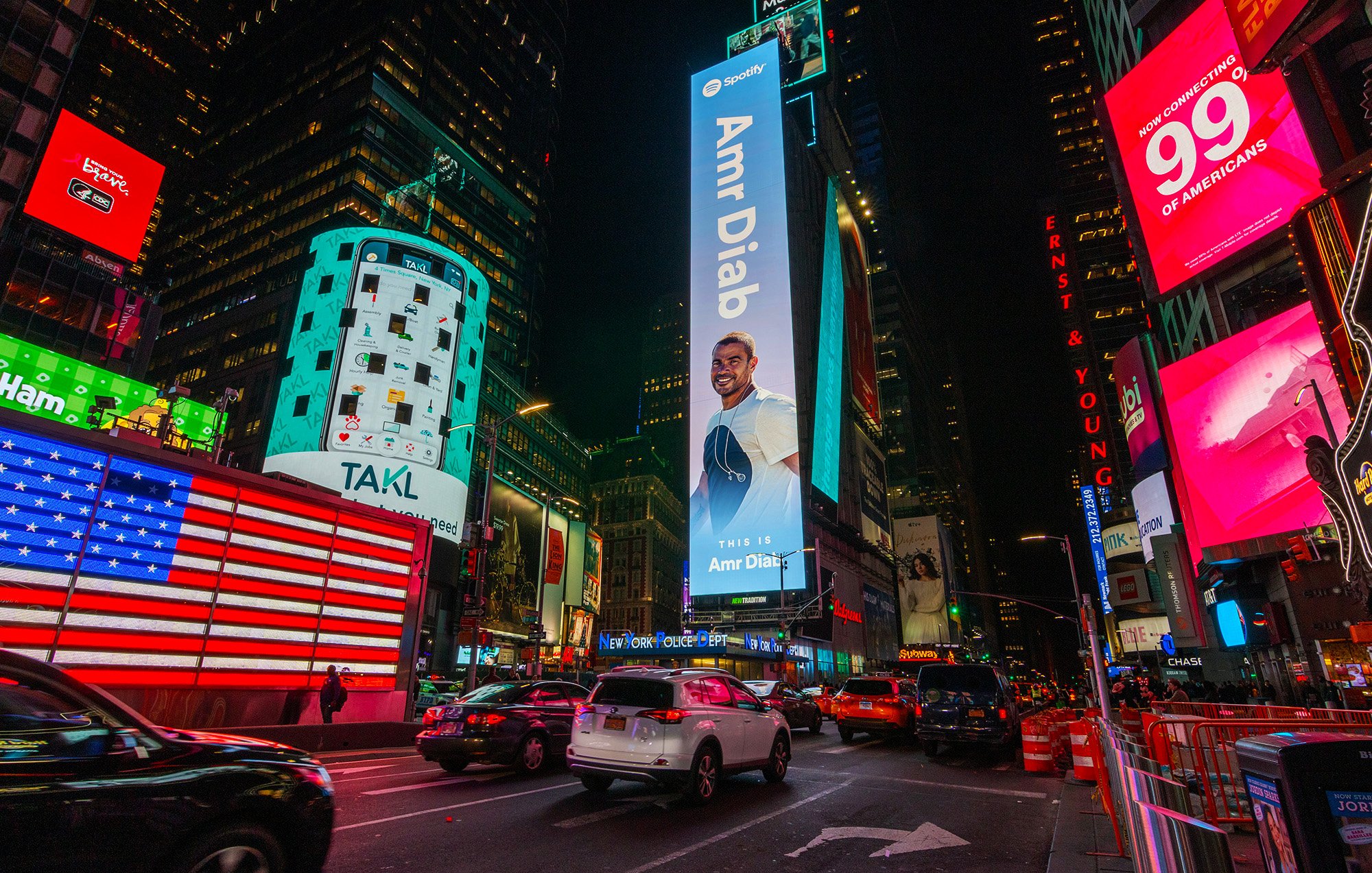 The image depicts a vibrant scene in Times Square, New York City, at night. The area is illuminated by numerous bright advertisements and digital billboards. Prominently featured is a large advertisement for Amr Diab, an acclaimed musician, which invites viewers to listen to his music. There is an American flag displayed on one of the buildings, and several vehicles are seen navigating the busy street. The atmosphere is lively, characteristic of this iconic urban location.
