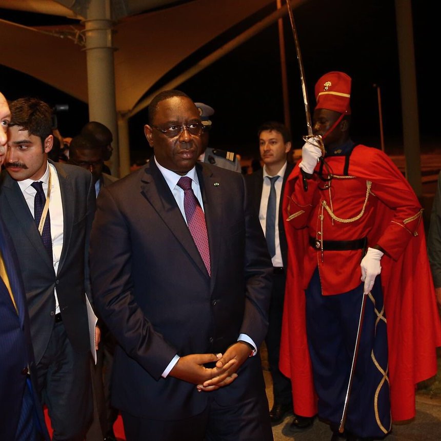 في الصورة، يظهر شخصان يرتديان بدلات رسمية، أحدهما هو الرئيس التركي رجب طيب أردوغان، والآخر هو رئيس دولة سنغافورة. يقف بجانبهم شخص آخر يرتدي بدلة. في الخلفية، توجد مجموعة من الأشخاص يرتدون ملابس رسمية أيضاً، بالإضافة إلى حارس يرتدي زياً تقليدياً. يبدو أنهم في موقع رسمي، ربما خلال زيارة أو استقبال رسمي. الاجواء تعكس الهدوء والدبلوماسية.