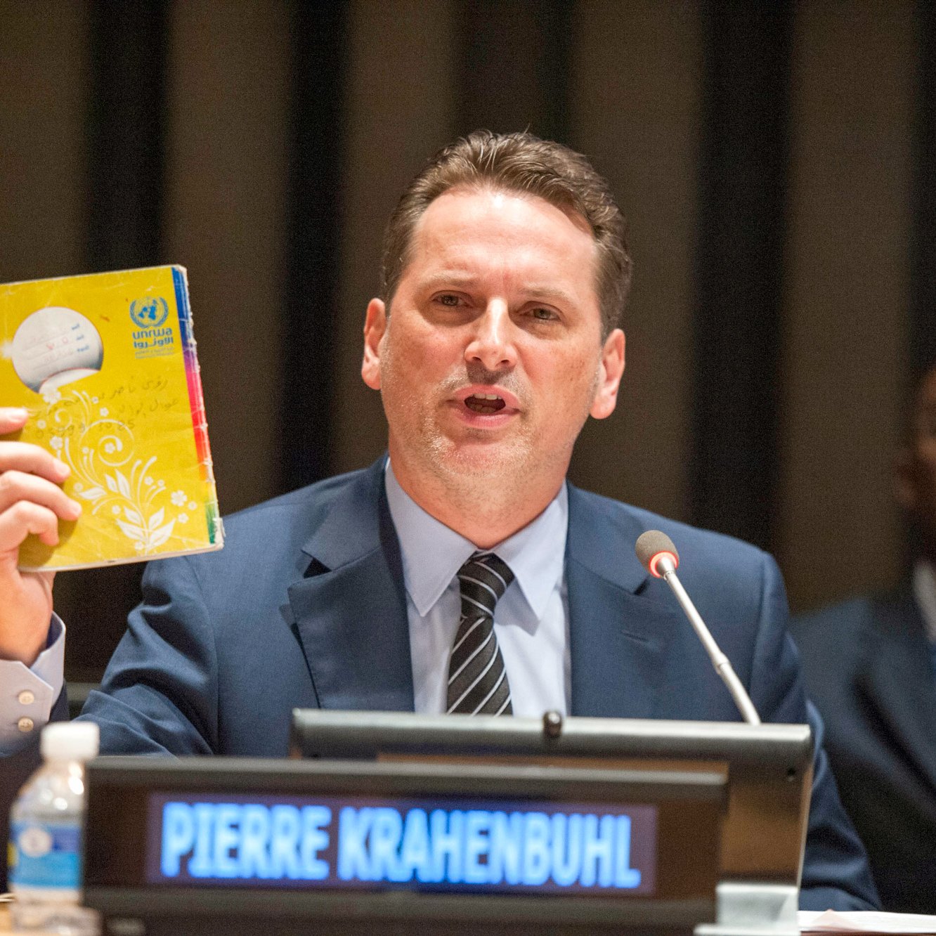 The image shows a man speaking at a podium during a conference or meeting, likely at a United Nations event. He is holding up a yellow booklet or document, which appears to be significant to his presentation. The nameplate in front of him reads "PIERRE KRAHENBUHL." In the background, there are several other individuals seated, and the setting has a formal atmosphere typical of international discussions. The man is engaged and emphasizing a point while addressing the audience.