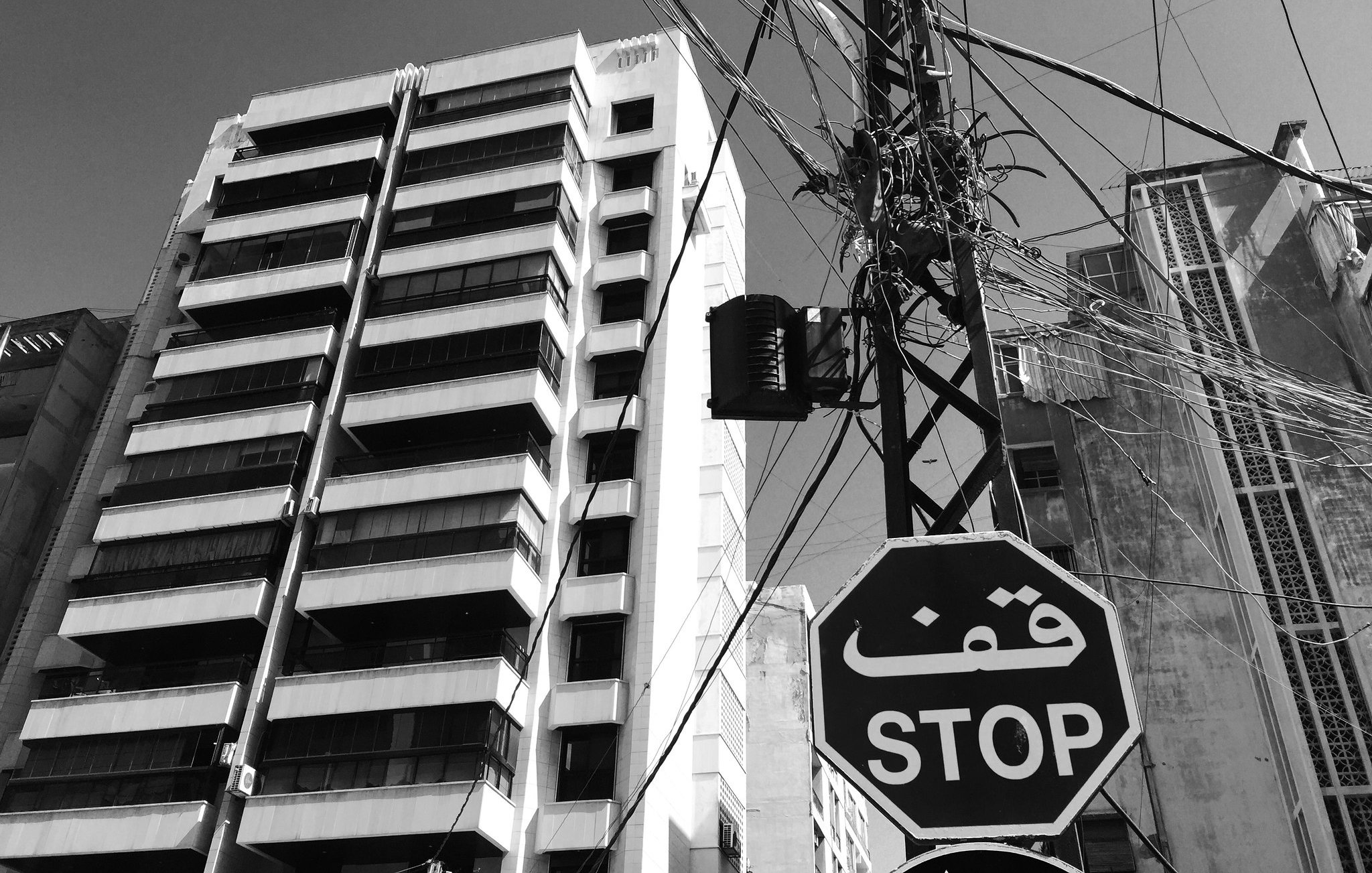 L'image montre une scène urbaine monochrome. À gauche, on aperçoit un bâtiment moderne avec plusieurs étages et balcons. À droite, un poteau électrique est visible, avec des fils enchevêtrés qui s'étendent dans le ciel. Il y a un panneau de signalisation en forme de hexagone avec le mot "STOP" écrit en arabe, ajouté à l'ambiance citadine. Le ciel est clair, ce qui donne une impression de luminosité à l'ensemble de la scène.