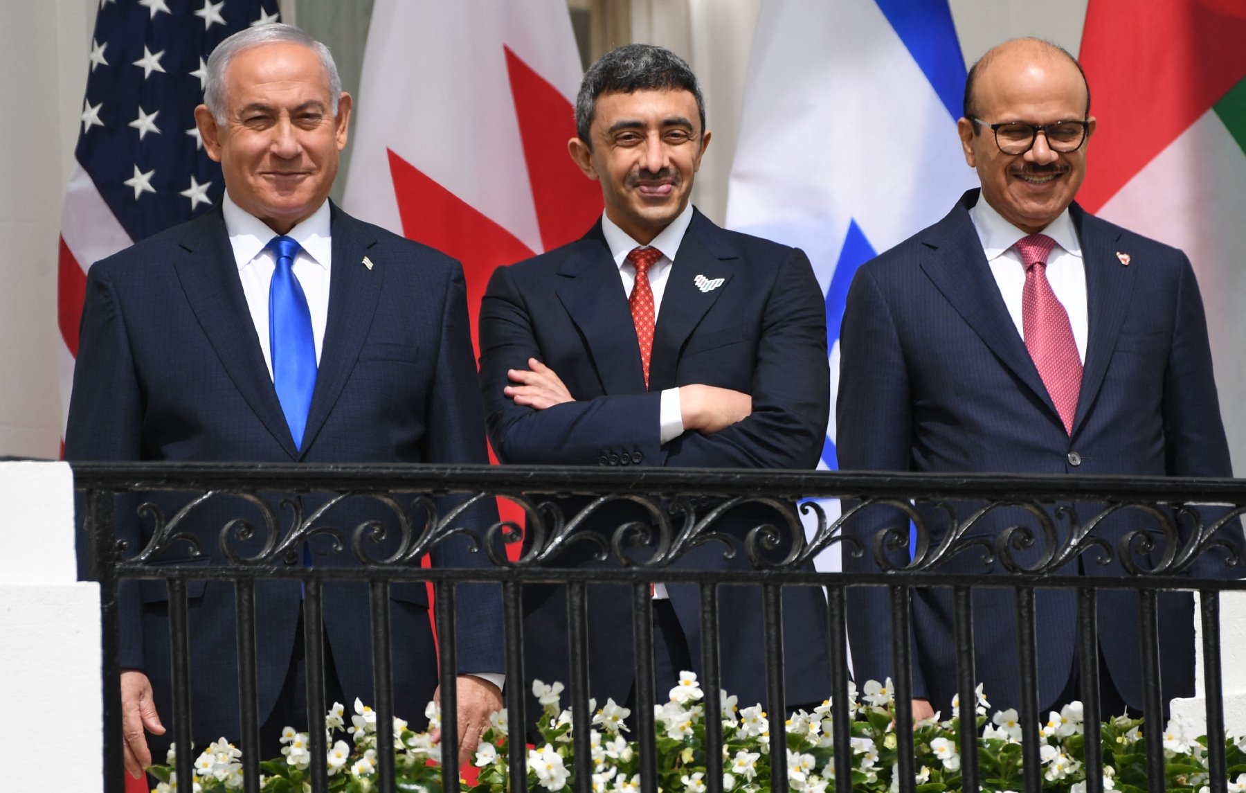 En la imagen se pueden ver a tres hombres de pie en una terraza, sonriendo. Al fondo hay banderas de Estados Unidos, Canadá, Israel y otros países. Los hombres están vestidos con trajes formales: el de la izquierda lleva una corbata azul, el del centro tiene una corbata roja y el de la derecha lleva una corbata a rayas rojas y azules. En la barandilla hay flores blancas. La atmósfera parece ser positiva y amistosa.