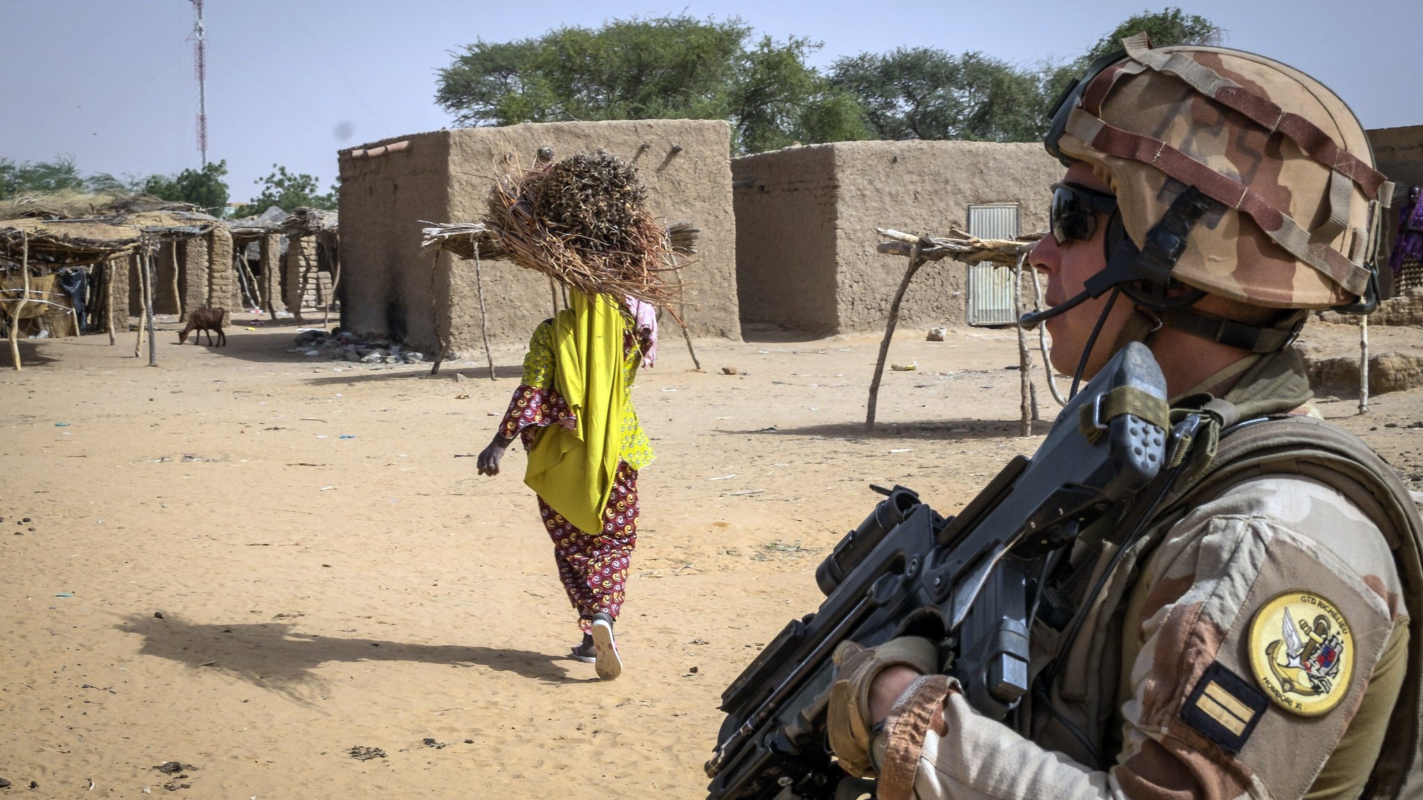 The image depicts a soldier standing in the foreground, holding a rifle and wearing military gear, including a helmet and camouflage uniform. The soldier appears vigilant as they face away from the camera. In the background, there is a woman walking away, carrying a large bundle of sticks on her head, dressed in a brightly colored traditional outfit. The setting looks like a rural area with simple mud-brick structures and some sparse vegetation, indicating a dry environment. The overall scene suggests a blend of military presence and civilian life in a remote location.